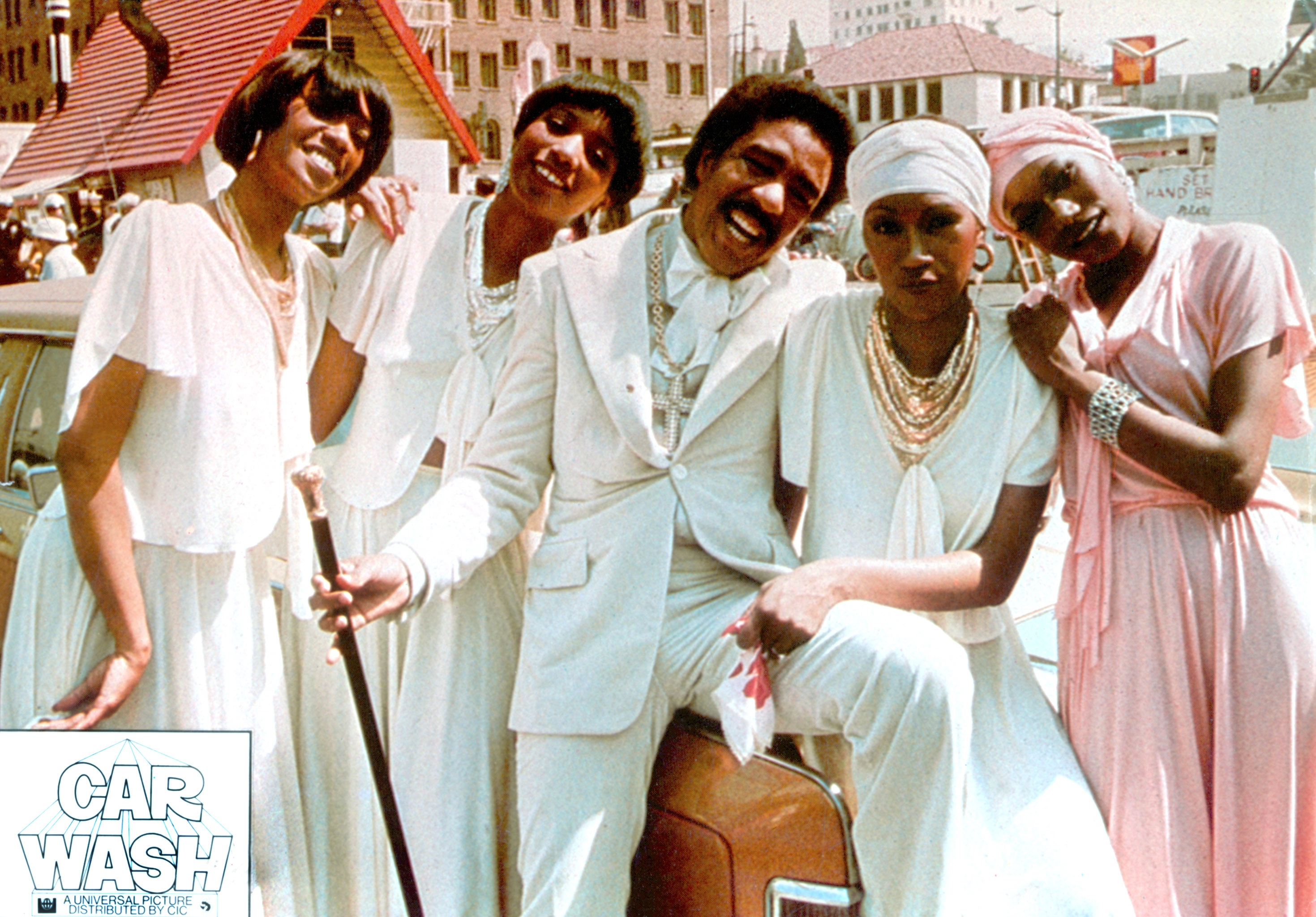 Five people in stylish 1970s attire pose cheerfully outside, leaning on a classic car, with a "Car Wash" movie title in the corner