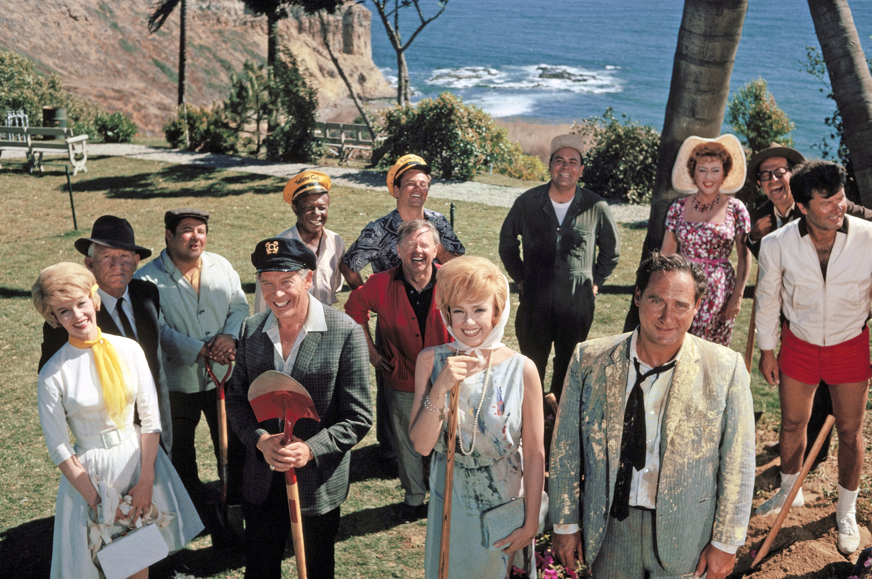 Group of people in eclectic 1960s attire, standing in a scenic outdoor area by the sea, possibly a cast photo from a TV show