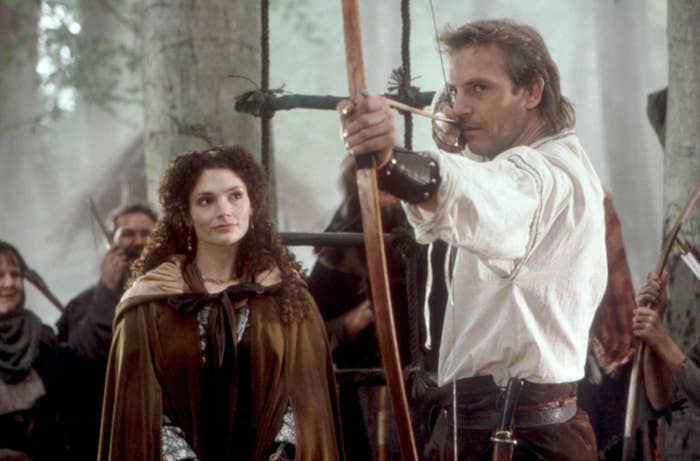 Actors portray medieval characters; one aims a bow and arrow, while another wears a cloak. Forest setting suggests a scene from a historical drama