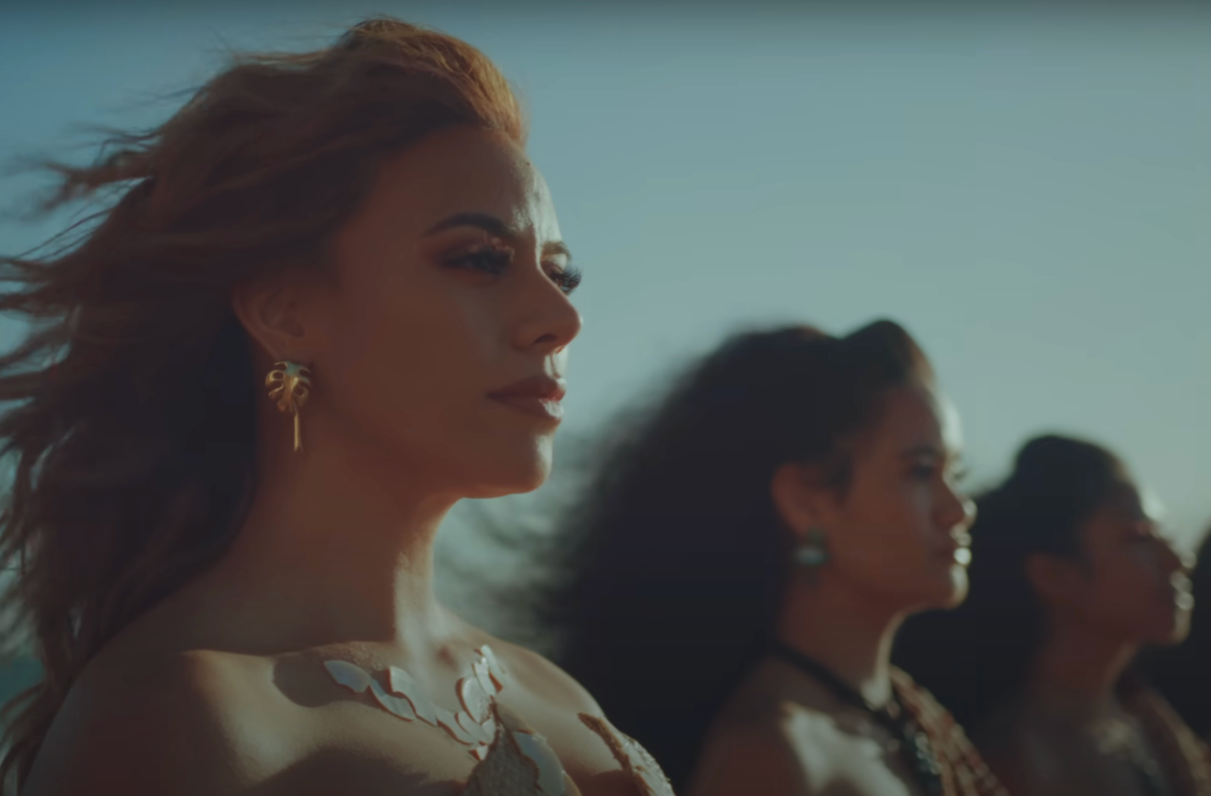 Three women face forward with serious expressions, wearing intricate jewelry and natural hairstyles, in a serene outdoor setting