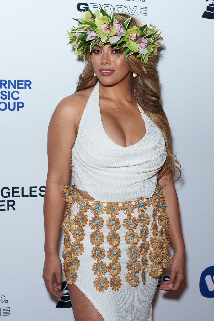 Person on red carpet wearing a white dress with a floral skirt overlay and a flower crown, smiling for the cameras