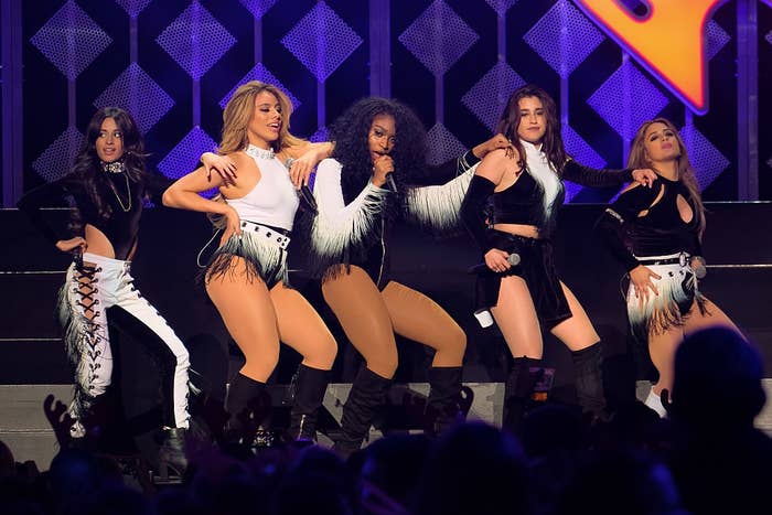 Five performers on stage wear coordinated fringe outfits, dancing energetically during a concert performance