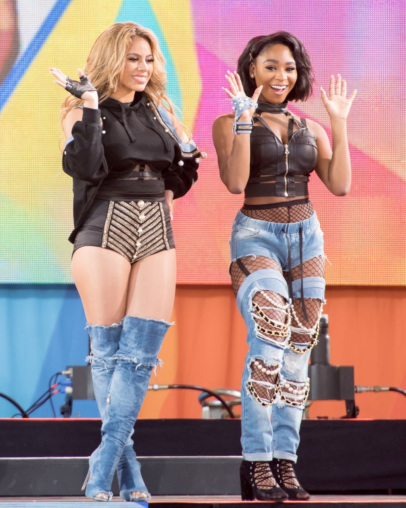 Two women posing on stage. One wears a black top, shorts, and denim boots. The other wears a leather top and patchwork jeans. Both are smiling and waving