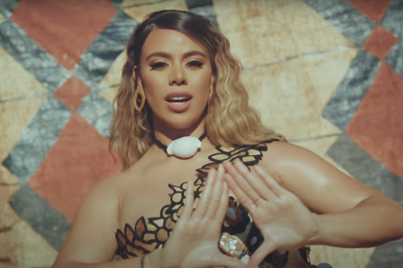 Person outdoors, wearing a patterned top with shell necklace, making a triangular hand gesture in front of a geometric background