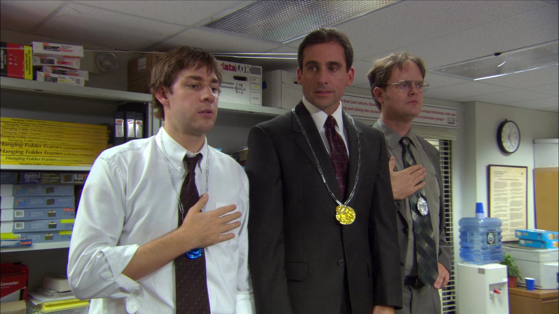 Three office workers stand solemnly with hands on hearts. One in a suit, others in shirts and ties, wearing fake medals in an office setting