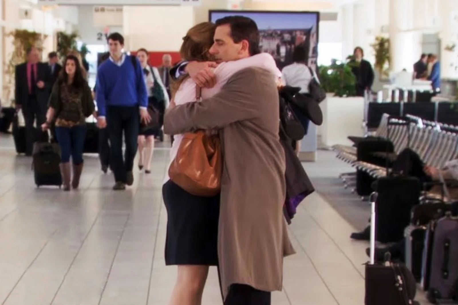 Two people embrace warmly in an airport setting, with travelers and luggage in the background