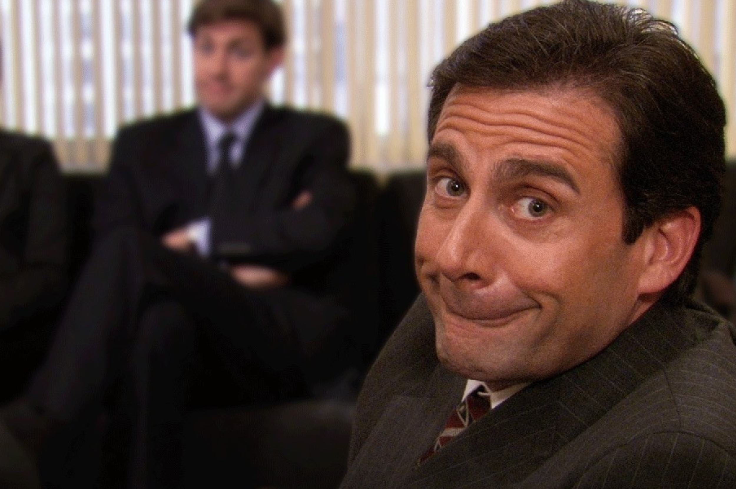 Man in a suit smirking at the camera; another man in a suit is blurred in the background, sitting on a couch