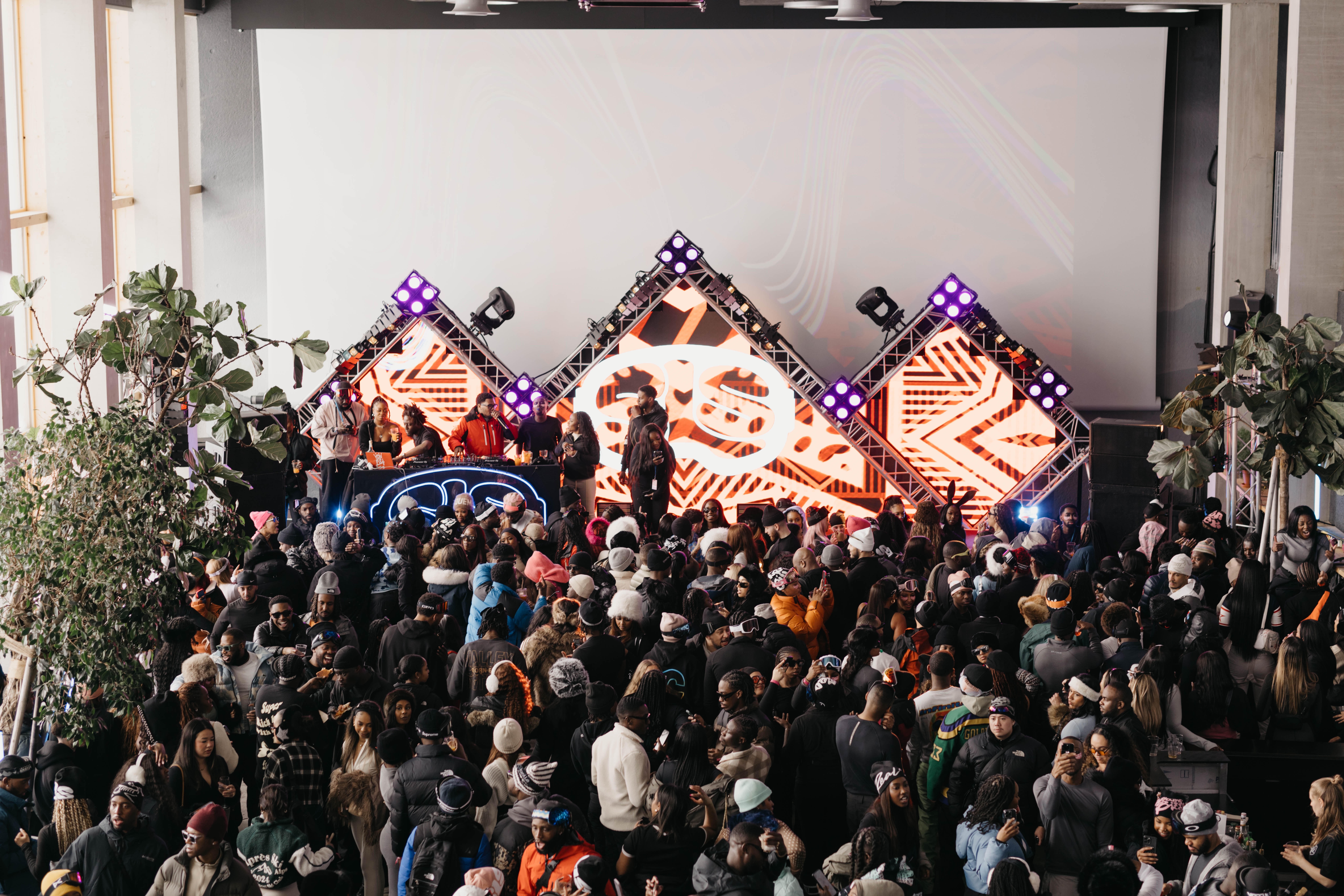 Large crowd at an indoor music event with a vibrant, geometric LED stage display and DJ performing