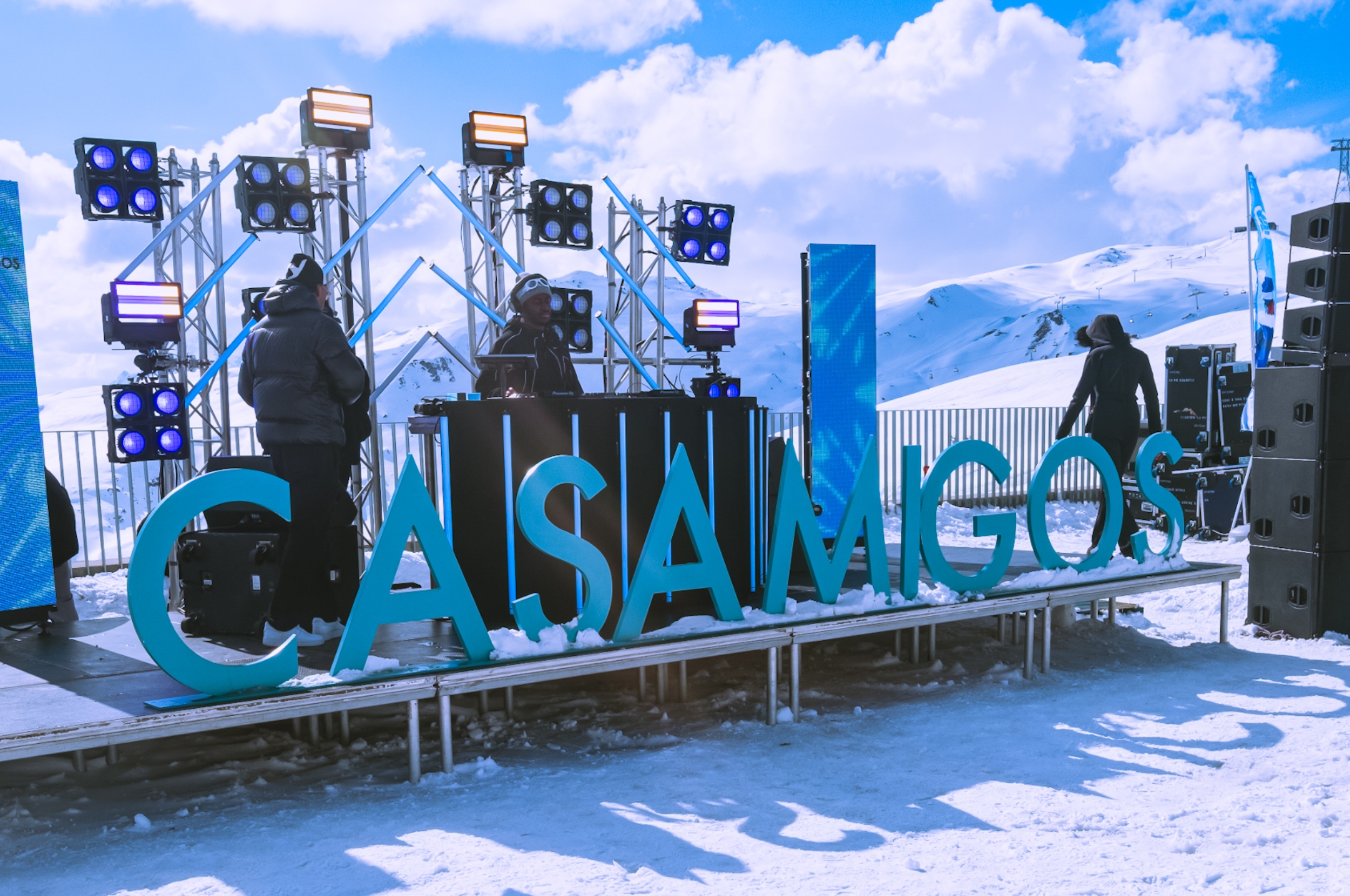 Outdoor DJ setup on a snowy mountain with large "CASAMIGOS" sign in front and two people on stage, surrounded by speakers and lights