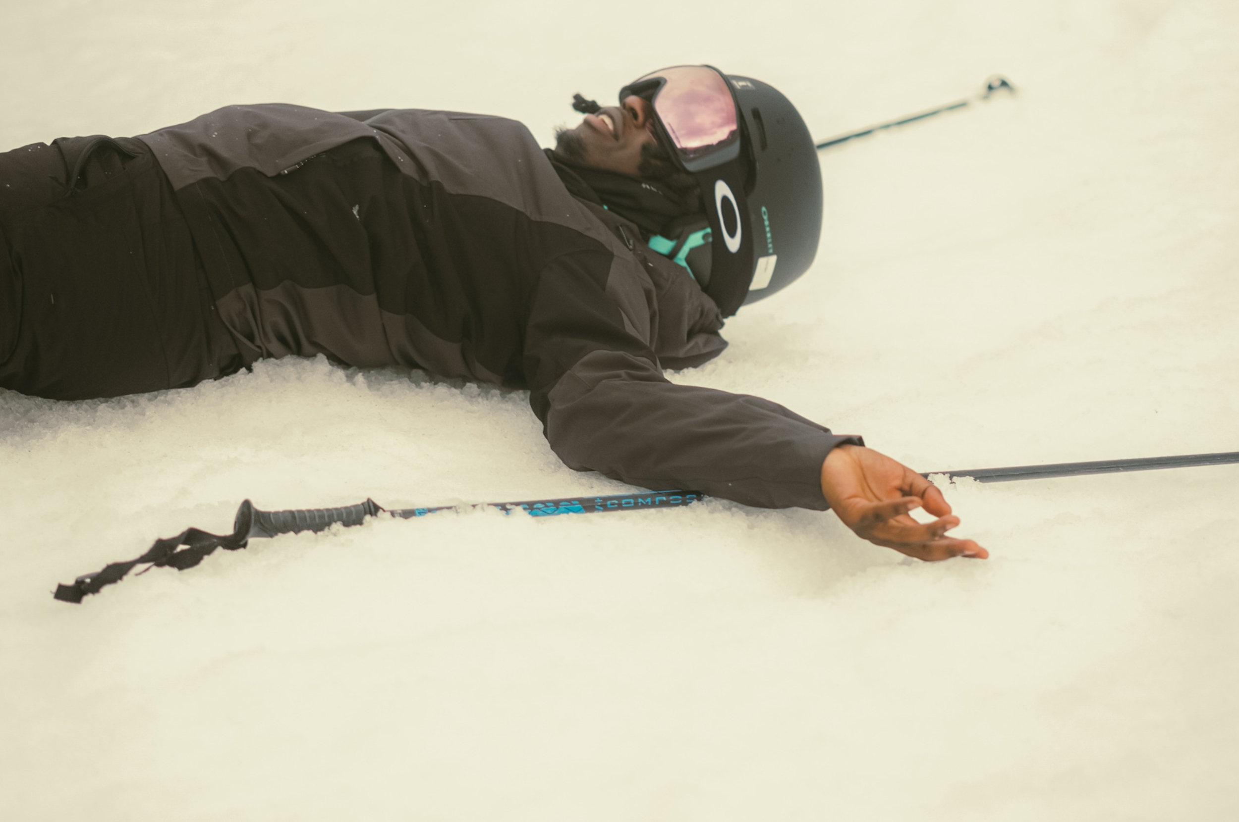 Person lying in the snow wearing ski gear, including a helmet and goggles, with skis beside them