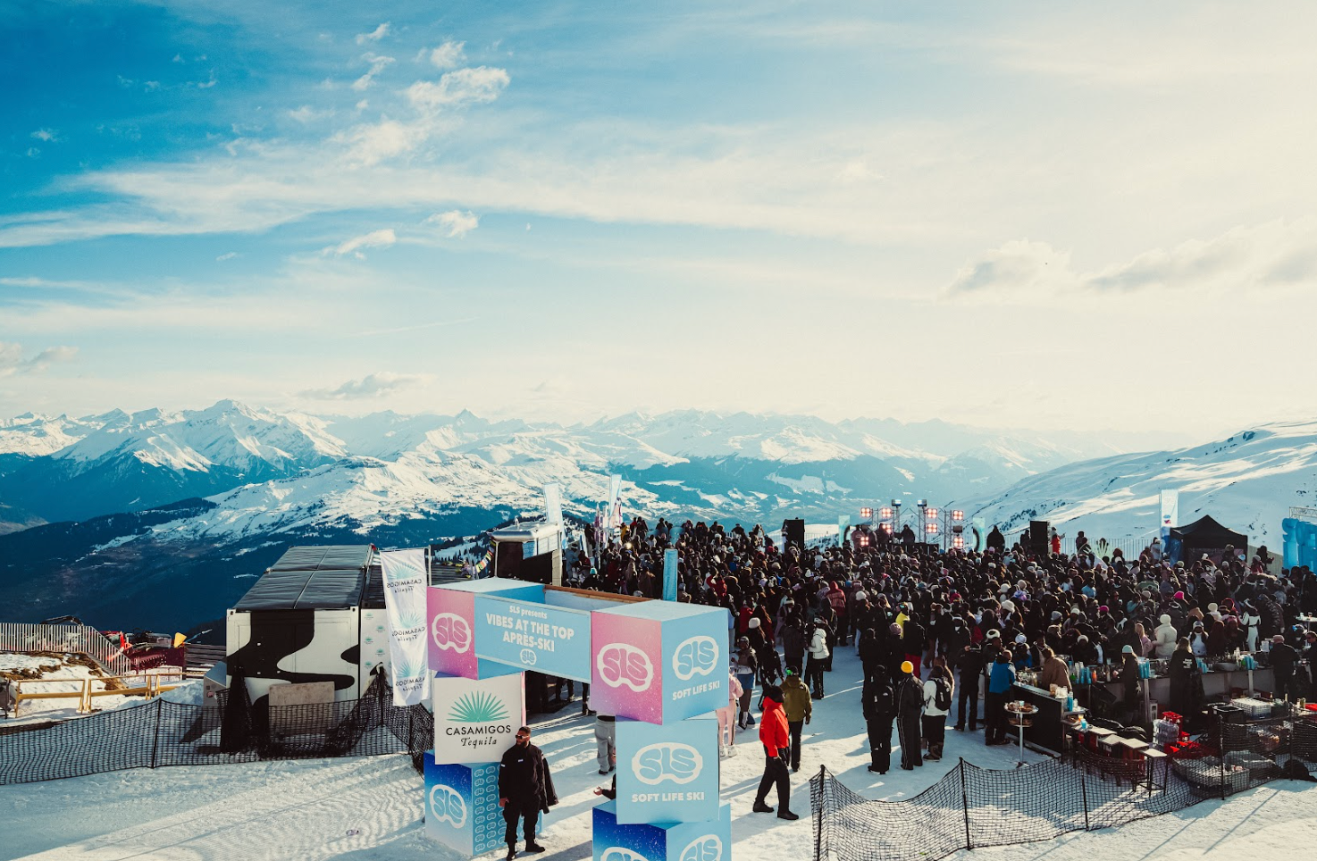 A crowd gathers at a snowy mountain festival with signs and booths, set against a backdrop of snow-covered peaks and a clear sky