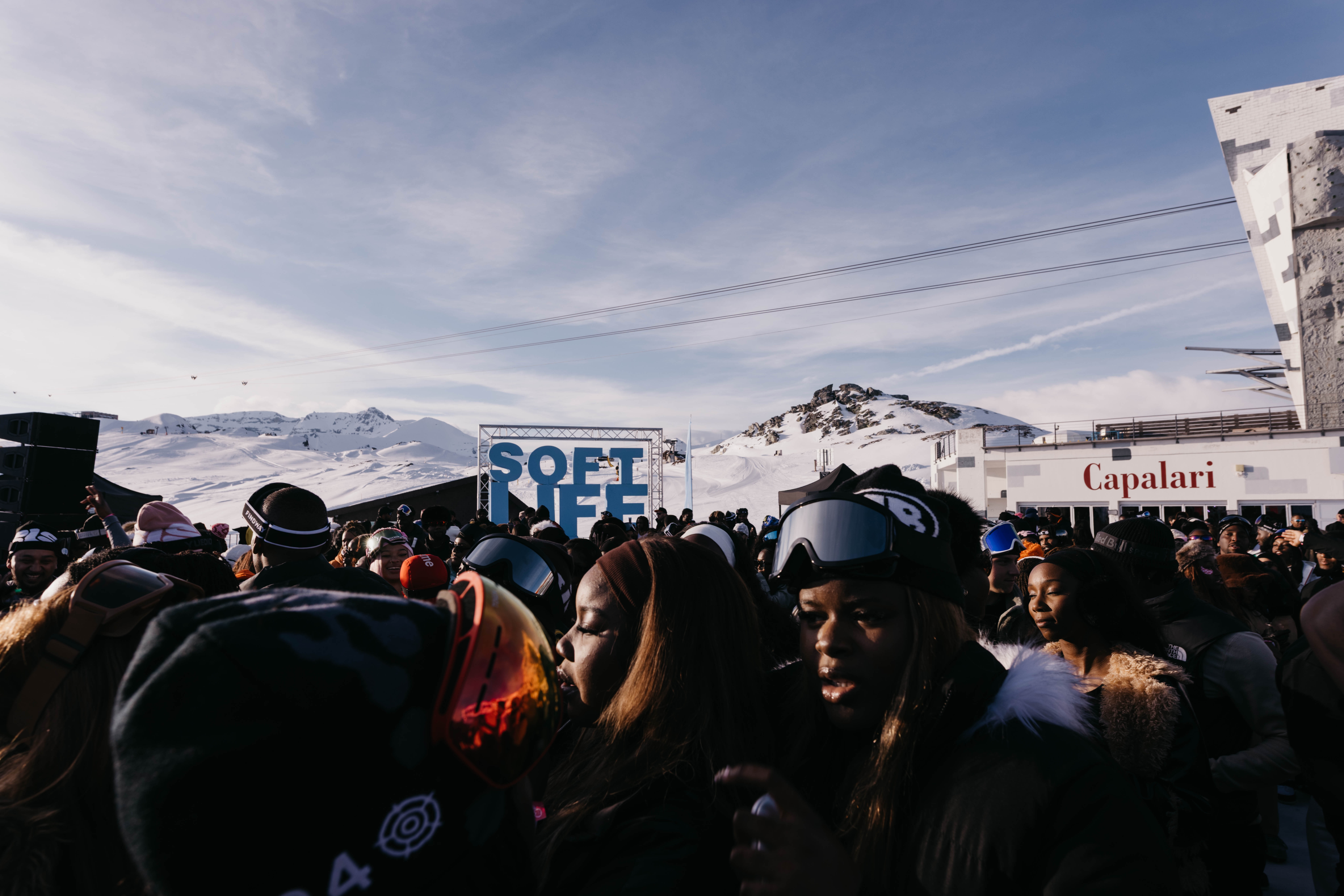 A crowd of people wearing winter gear and ski goggles at a snowy mountain resort event with a "Soft Life" sign in the background