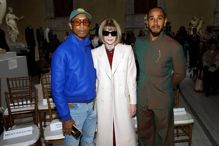 Three individuals pose at a fashion event. Two wear modern casual attire; the third wears a white coat with sunglasses. There are empty chairs behind them