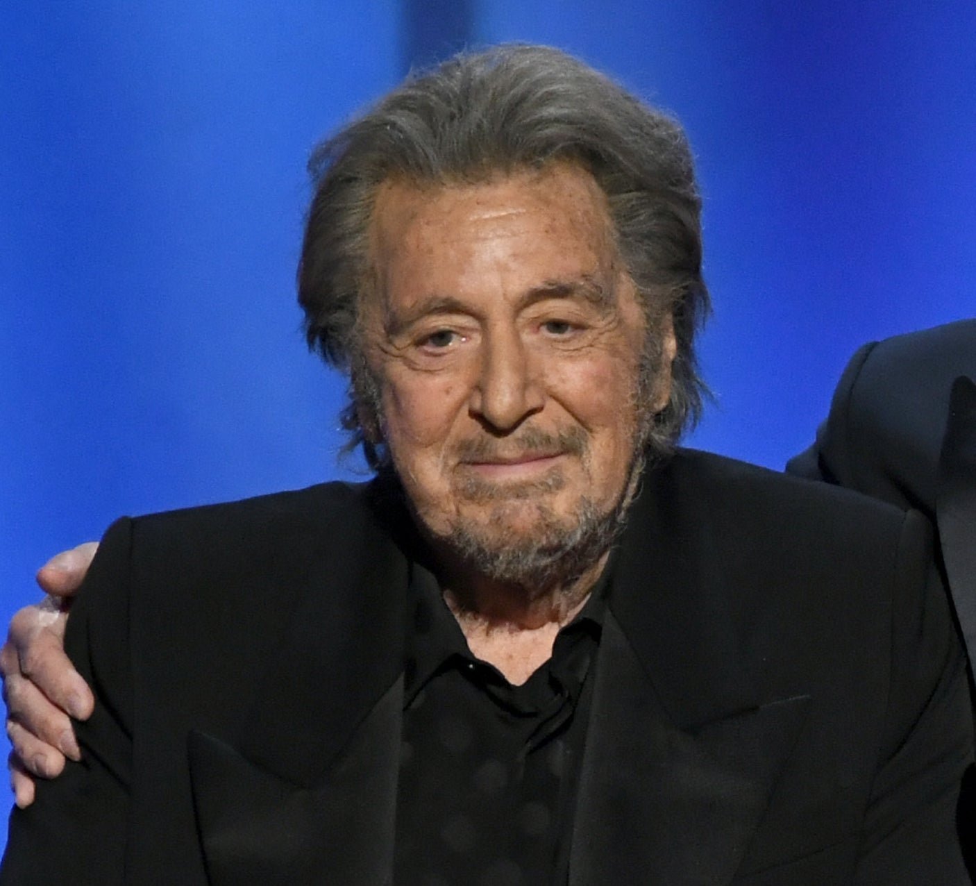 Two well-dressed men stand together on stage at an AFI event, smiling and embracing, in front of a podium with a microphone
