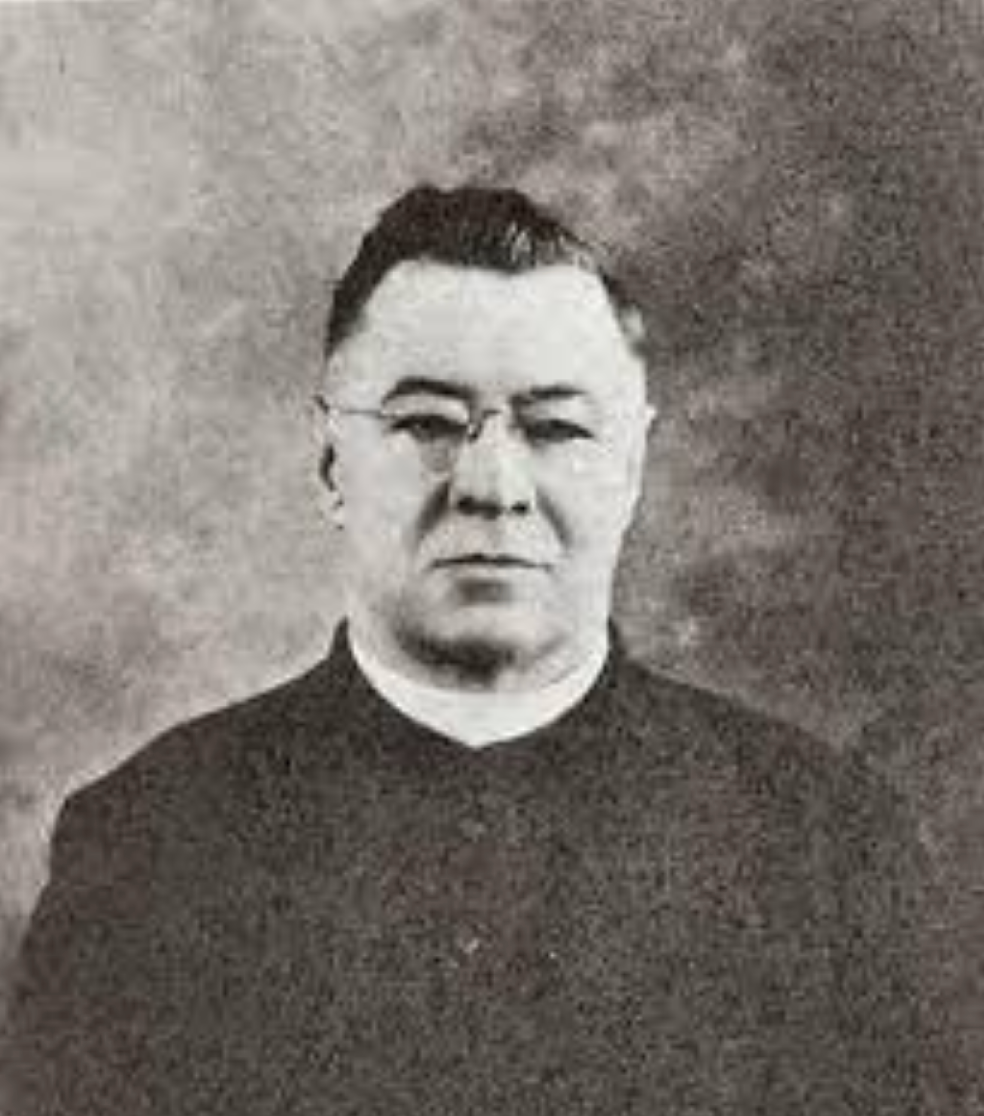 An unidentified man with glasses, wearing a dark suit, poses for a formal portrait against a textured backdrop