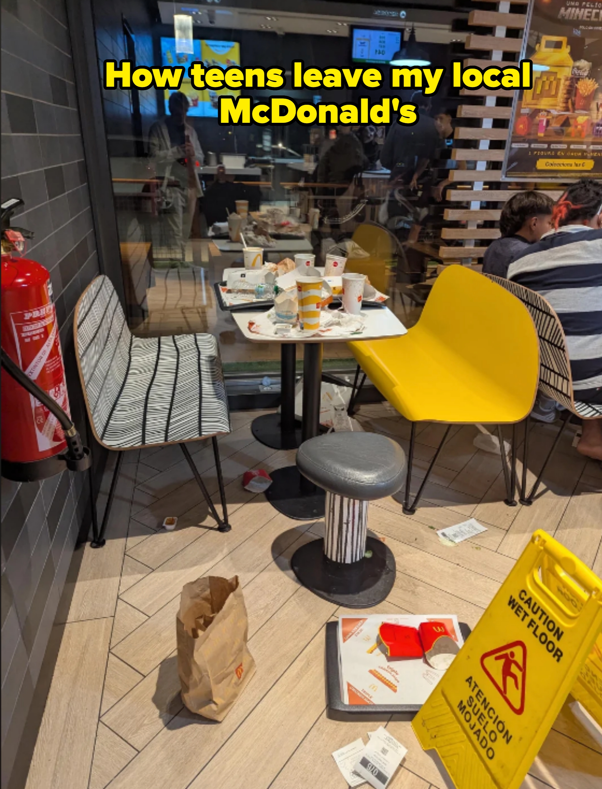 Messy fast-food restaurant table with empty food containers, chairs, and a wet floor sign nearby. Caption: &quot;How teens leave my local McDonald&#x27;s&quot;