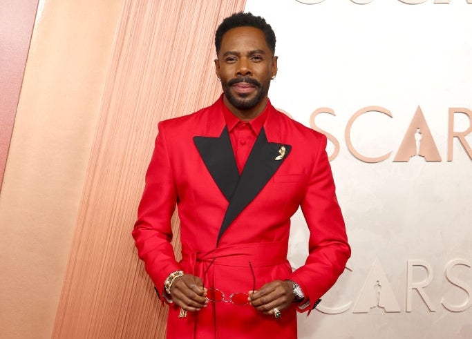 Person in a bold red and black tailored suit with a tie belt, standing on the red carpet at an event with "OSCARS" in the background