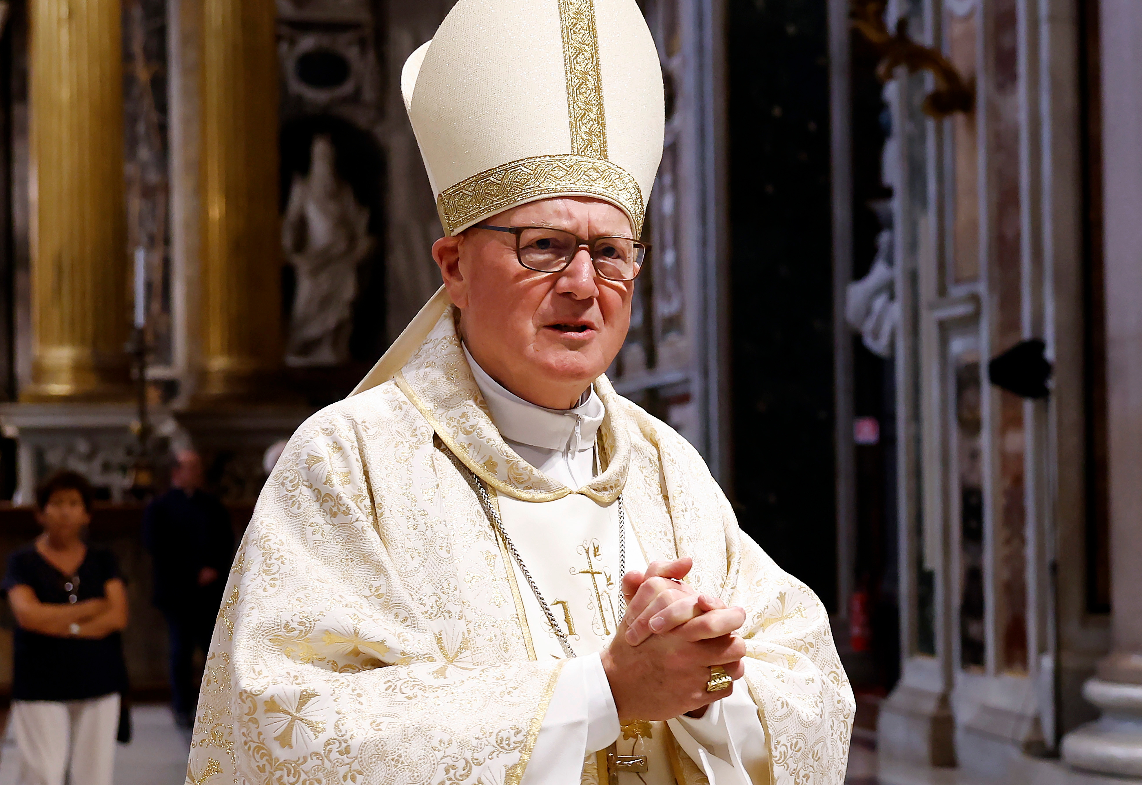 A religious leader in ornate ceremonial robes and a miter stands in an ornate interior, seen clasping hands during an event