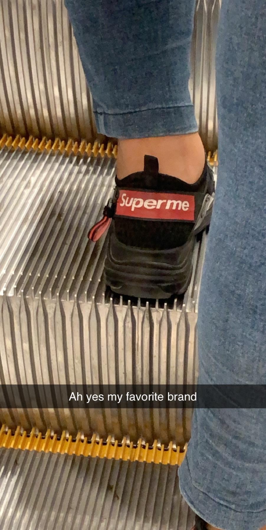 Close-up of a shoe on an escalator with a red and white label reading "Superme." Caption reads, "Ah yes my favorite brand."