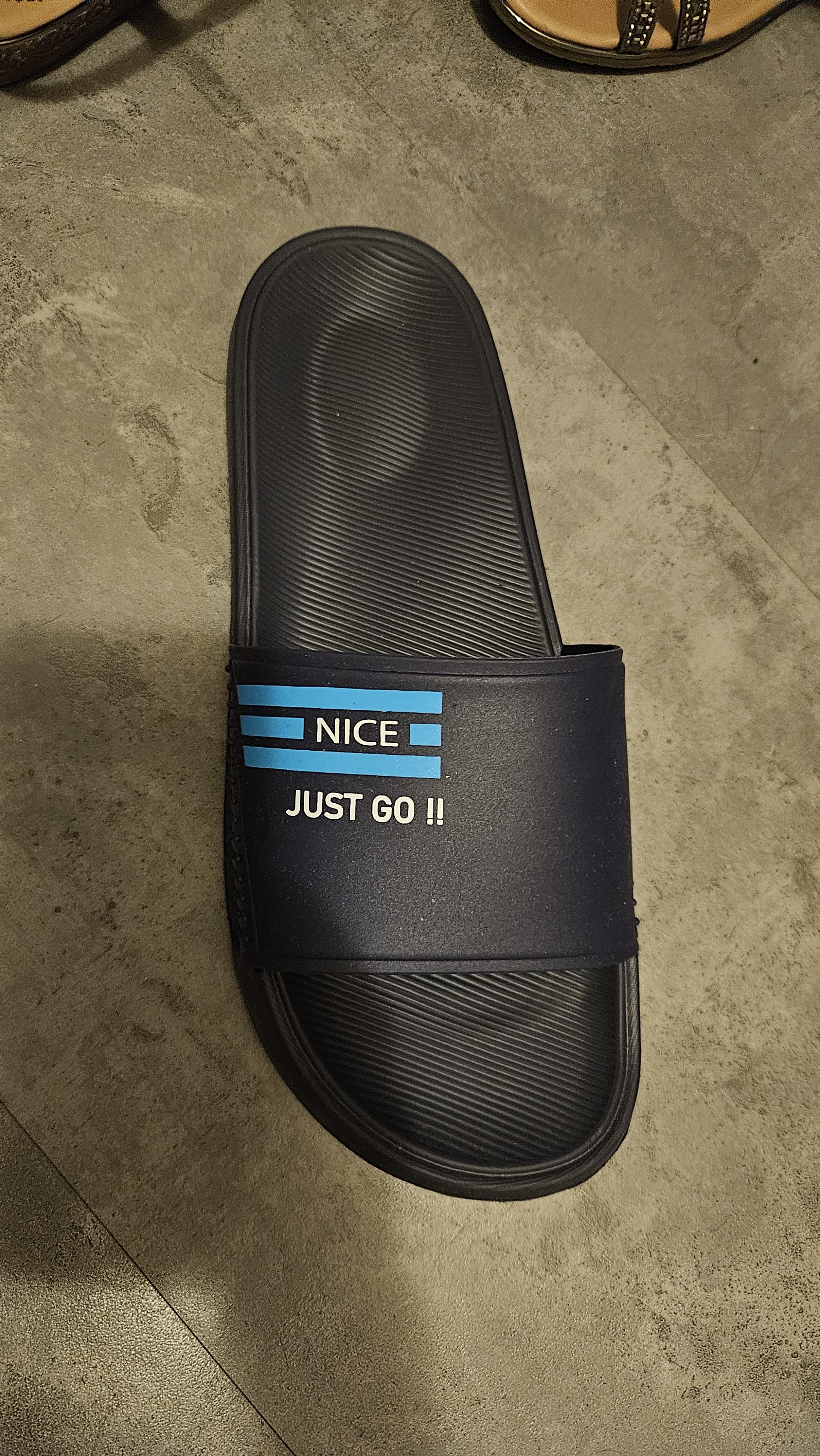 A single black slide sandal on a floor, with text "NICE JUST GO !!" on the strap