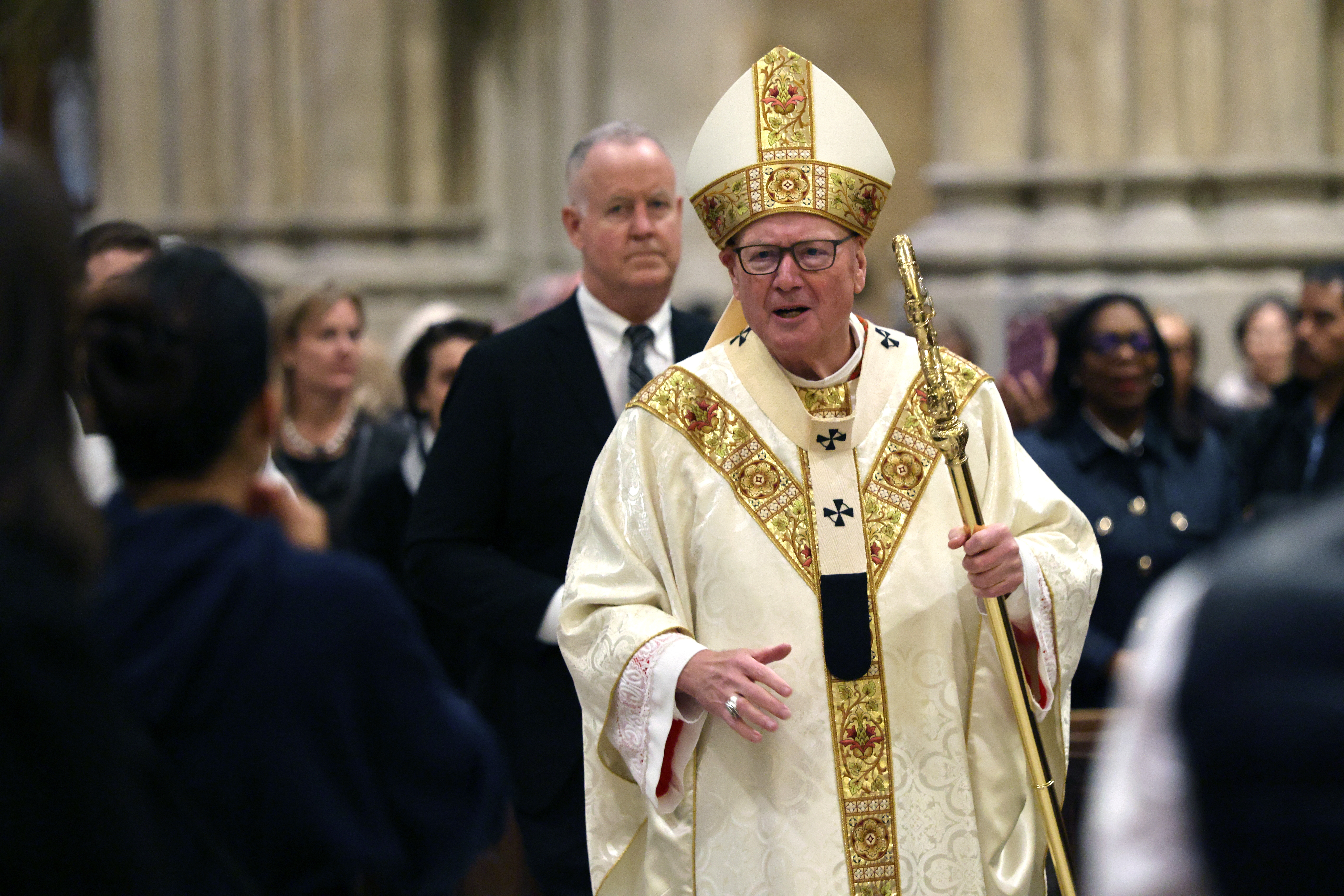 A religious figure in ornate ceremonial attire walks in a procession, holding a staff, with people and a grand interior in the background