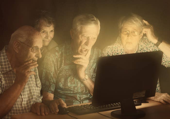Four older adults are gathered around a glowing computer screen, appearing intrigued and surprised by what they see
