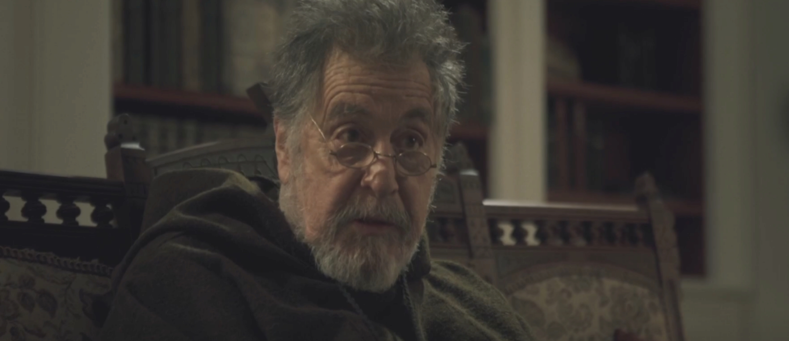 Elderly man with glasses and a hooded coat sits in a dimly lit room, looking pensive