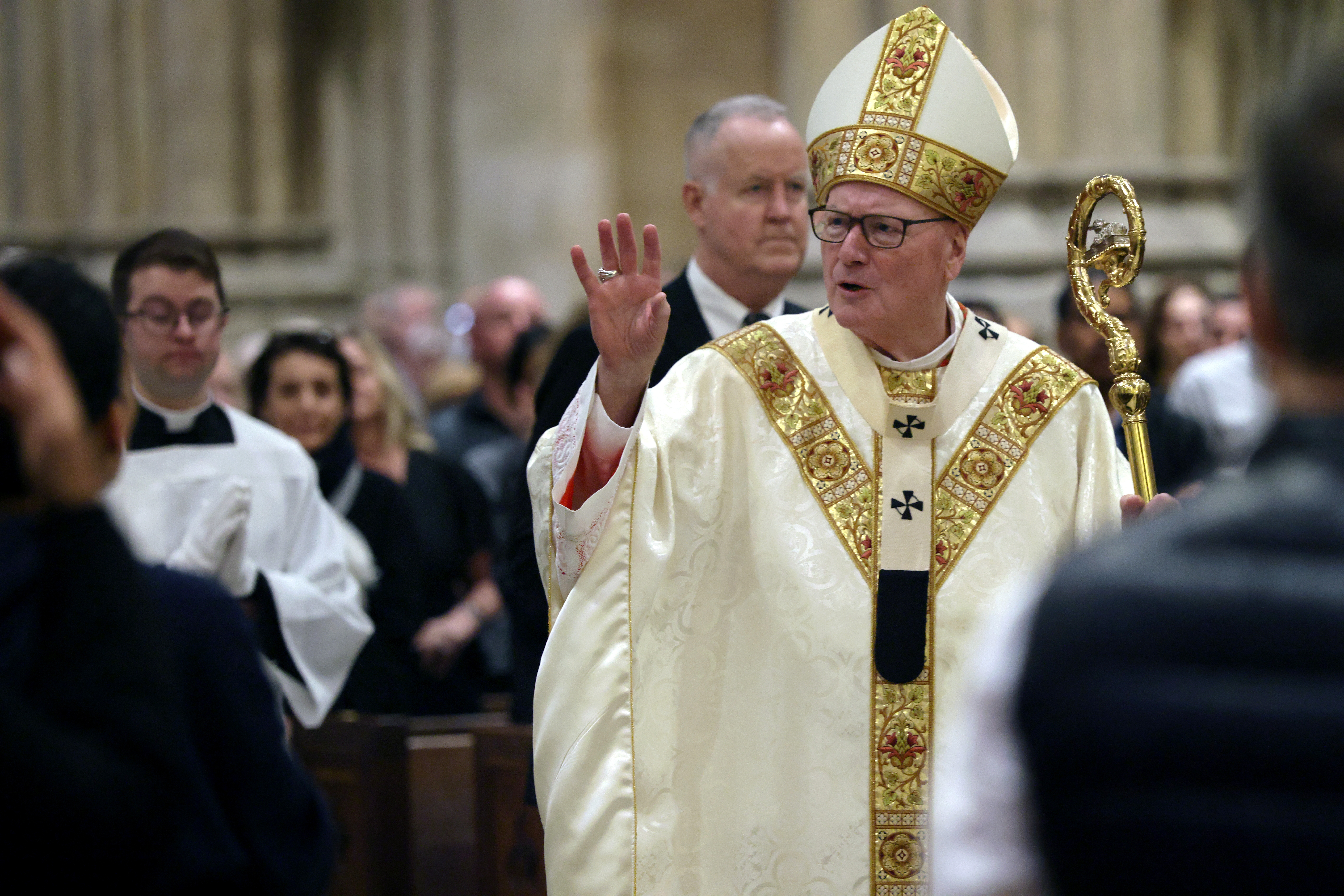 A religious leader in ornate ceremonial attire raises a hand in blessing during a church service, surrounded by attendees