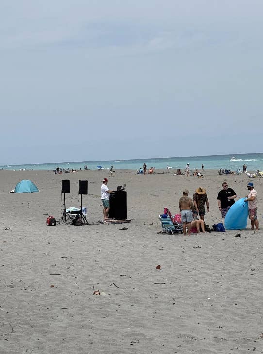 Person on a beach with a laptop and speakers set up; nearby people relax and chat, enjoying a day by the sea