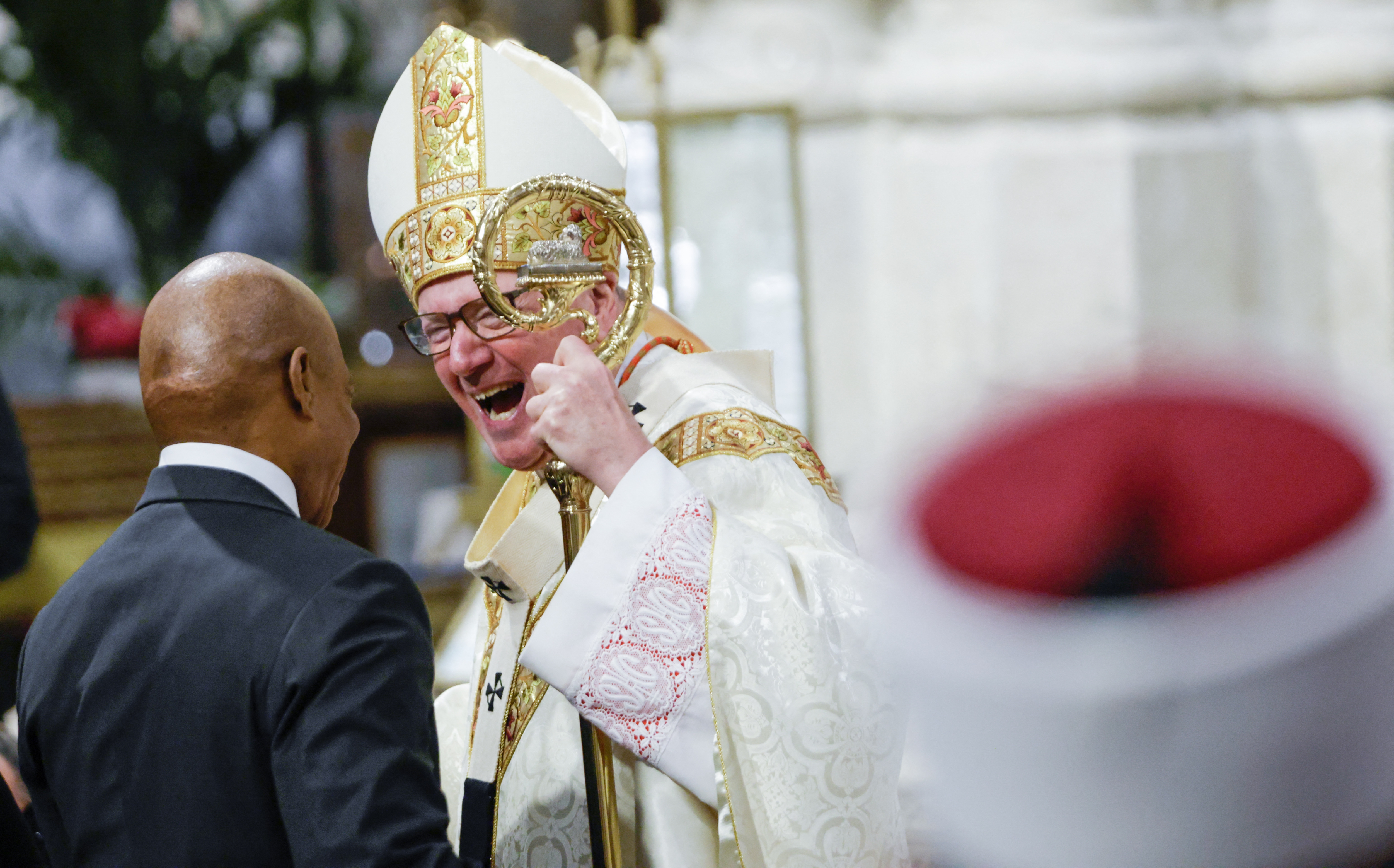 A high-ranking clergyman in ornate religious robes smiles and gestures while speaking with a person in a suit at a formal event