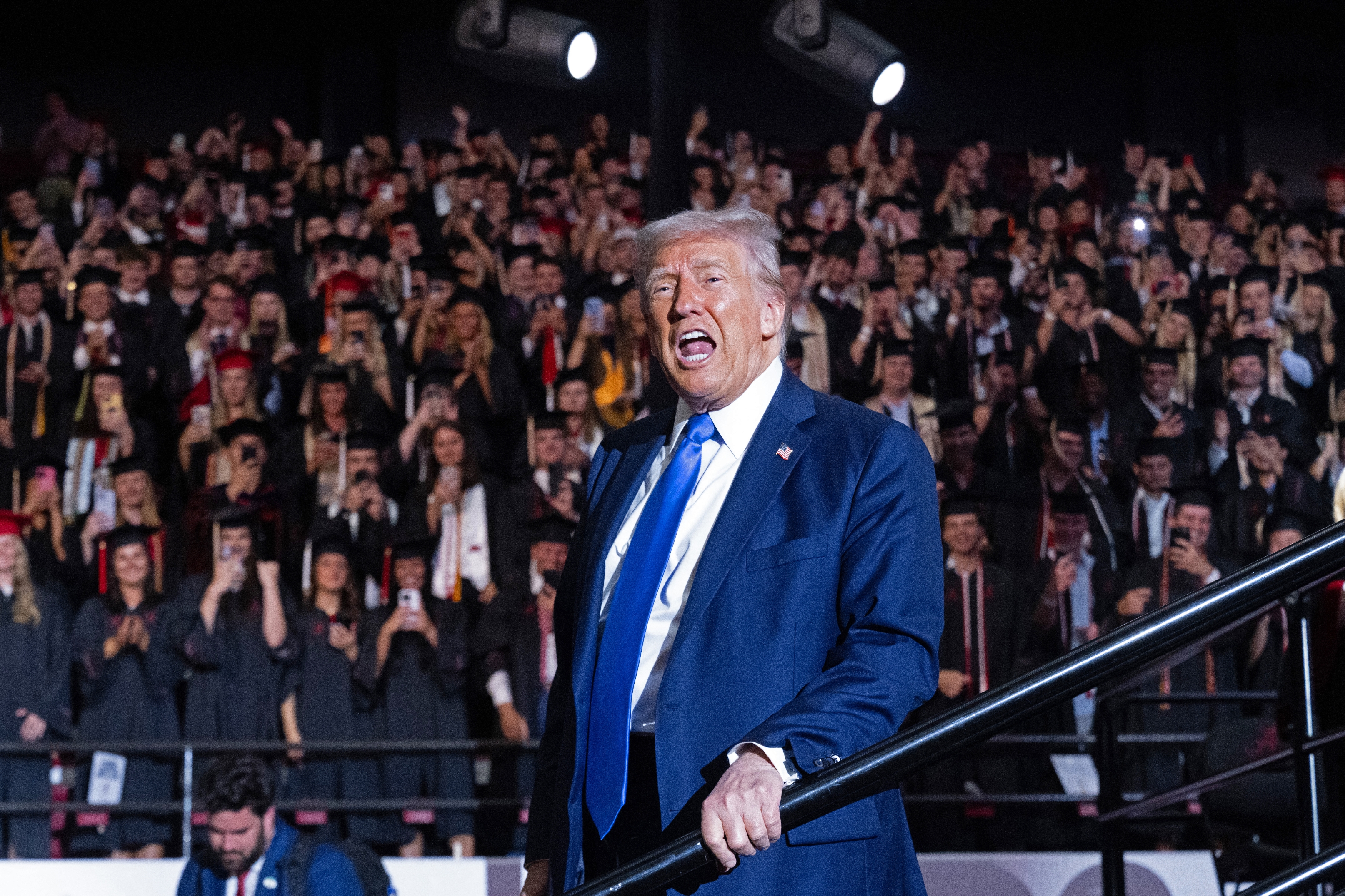 Individual in a suit speaks energetically on stage at a graduation event, with a crowd of applauding graduates in the background