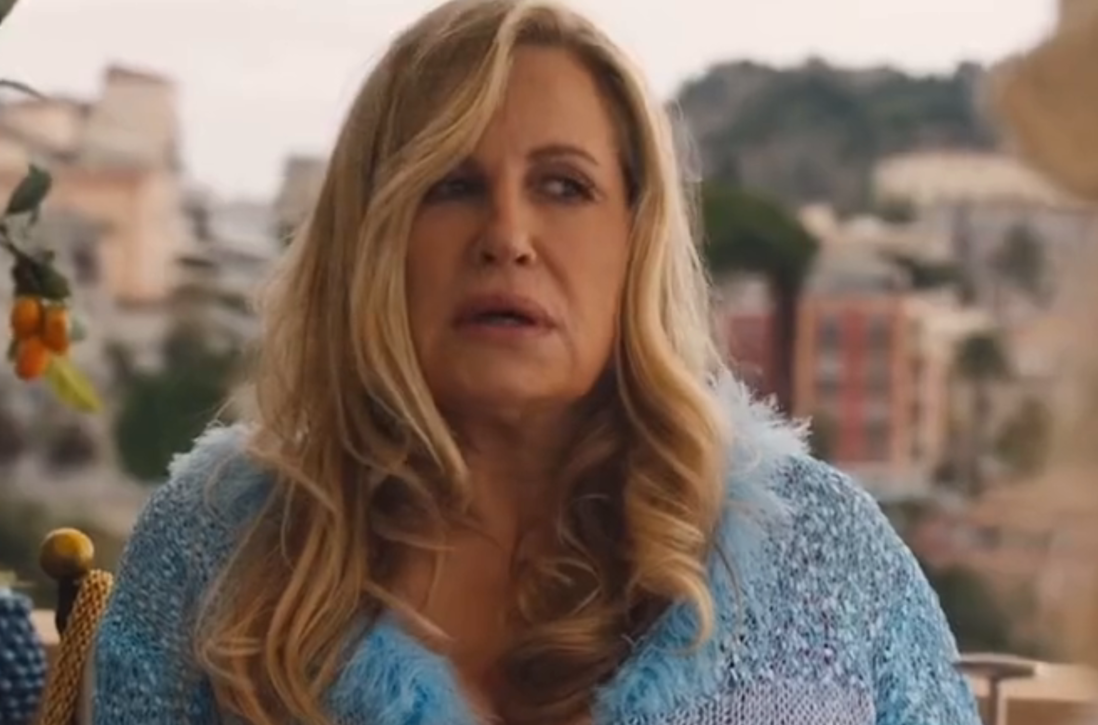 Woman with long, wavy hair in a textured outfit giving an expressive look, seated outdoors with blurred buildings in the background