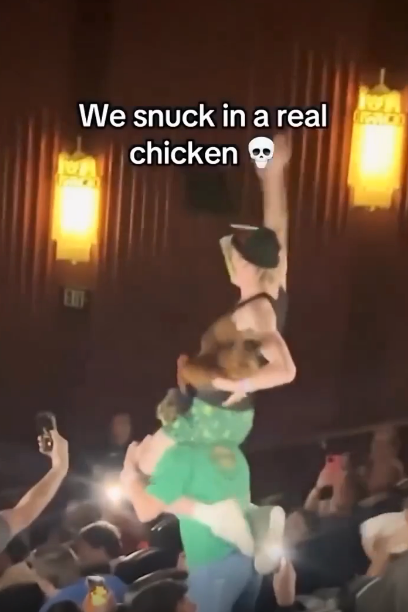 Person on another&#x27;s shoulders holding chicken inside theater; audience watches, some with phones. Text reads: &quot;We snuck in a real chicken&quot;