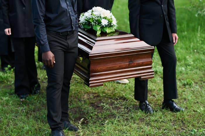 People carrying a wooden casket adorned with white flowers at an outdoor ceremony