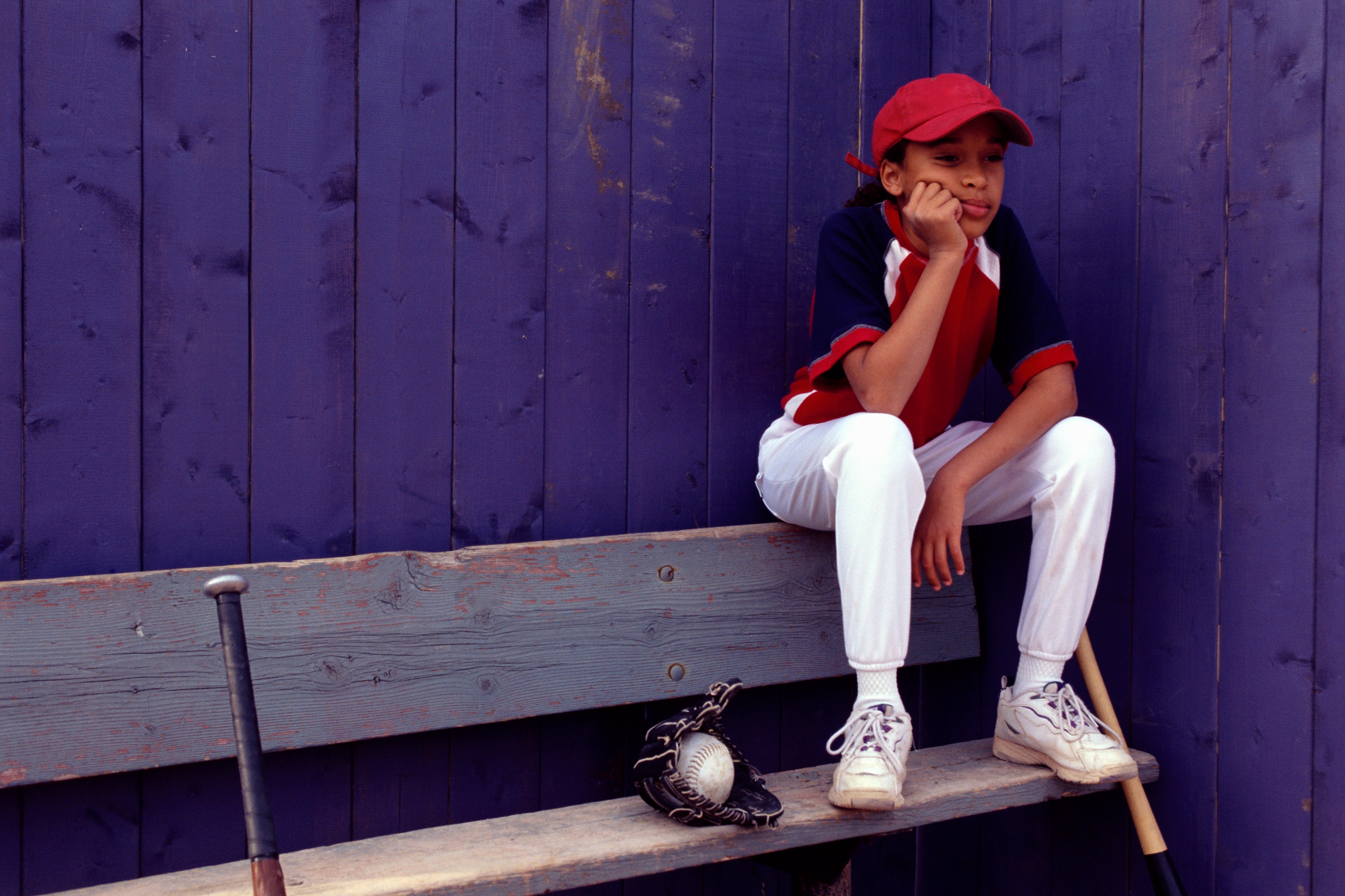Child in baseball uniform sitting on a bench, resting their head on their hand, with a glove, ball, and bat nearby
