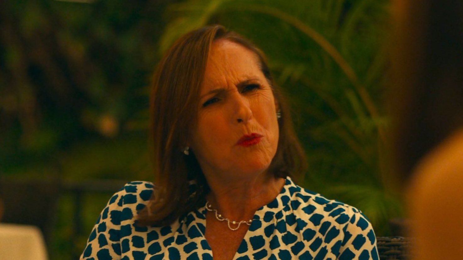 Woman with shoulder-length hair looks confused, wearing a patterned top and necklace. She's seated outdoors, with foliage in the background
