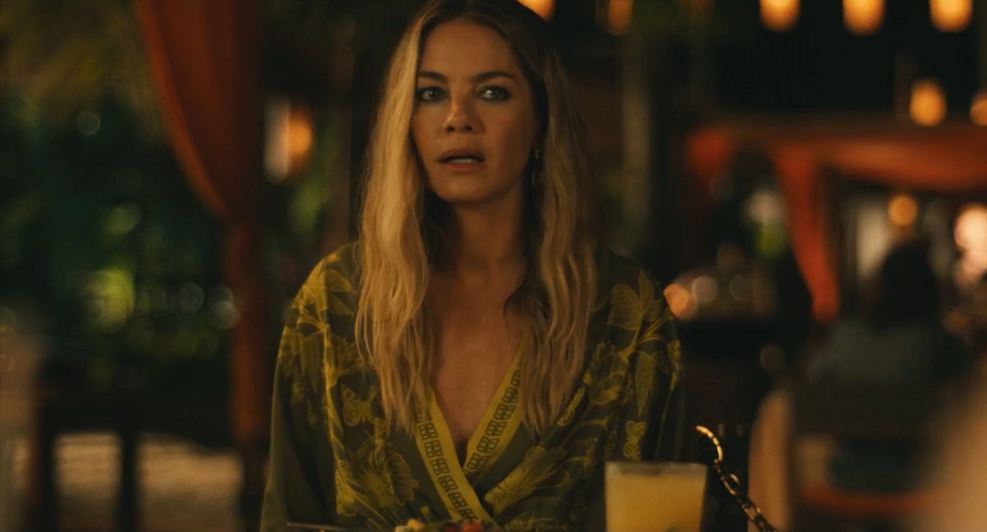 Woman with long hair in a printed dress looking surprised, seated at a dimly lit restaurant table with a drink in front of her