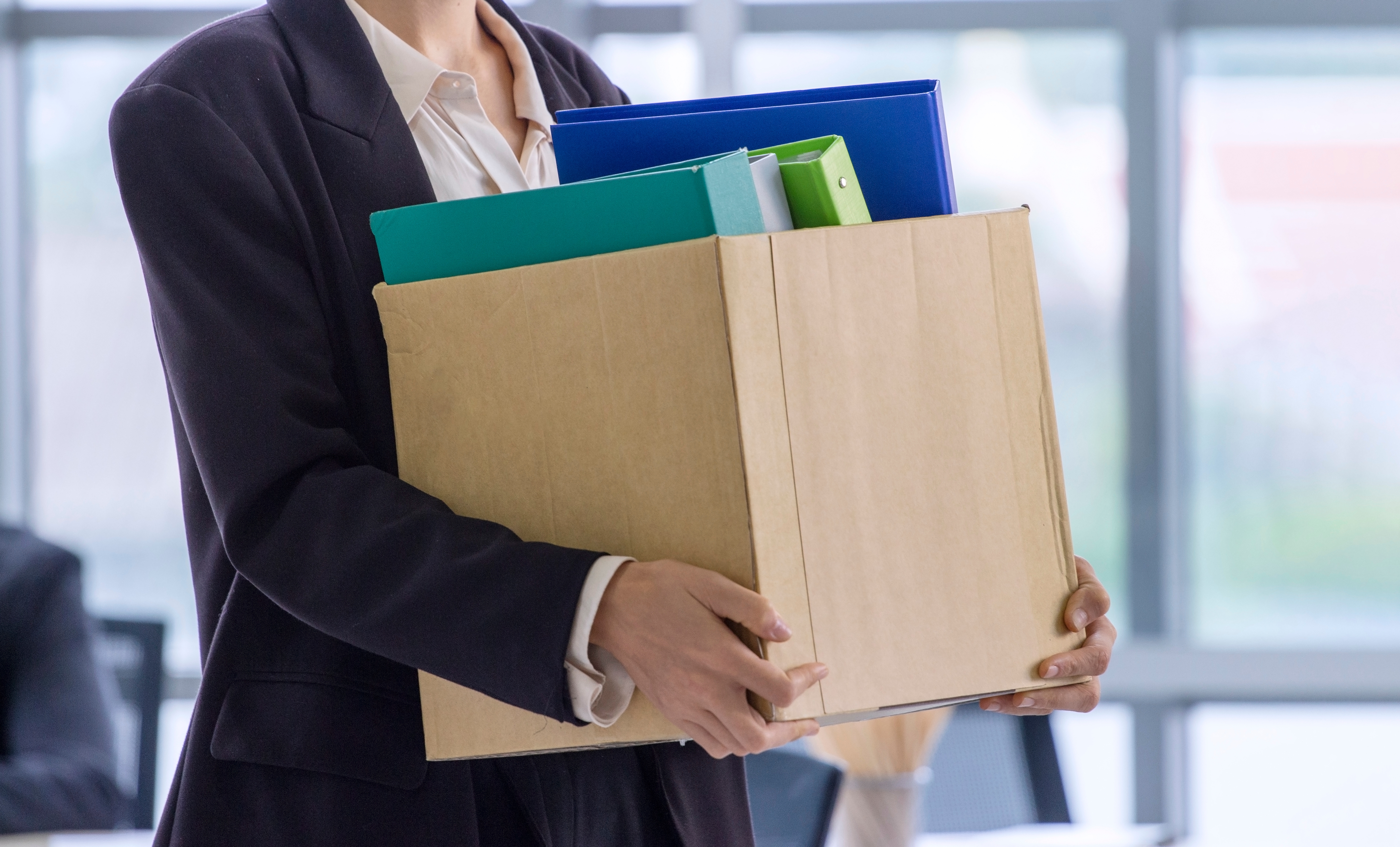 Person in a suit holding a cardboard box filled with binders and files, hinting at an office move or job transition