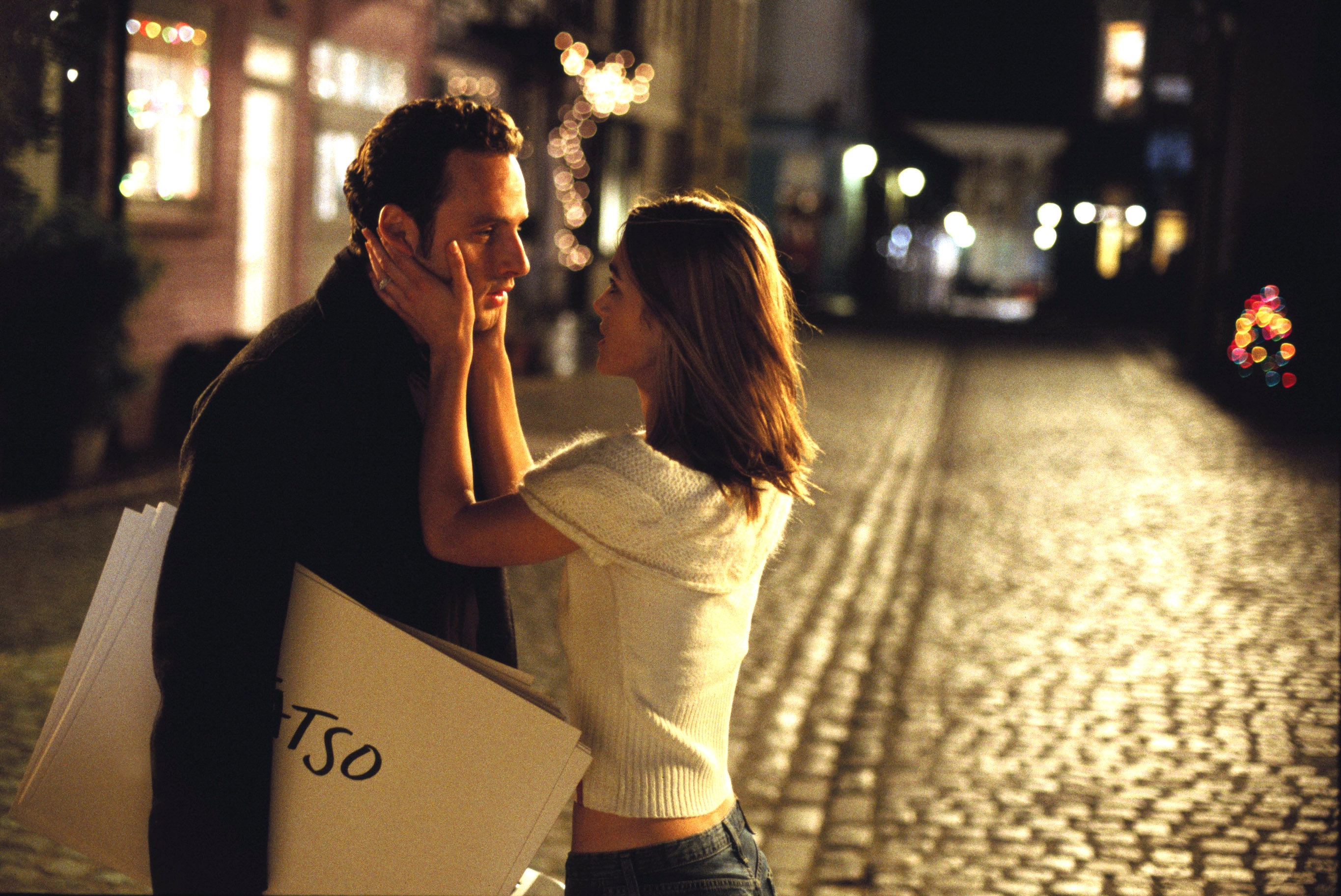 A romantic scene at night on a cobblestone street. A woman gently holds a man's face; he holds large cards
