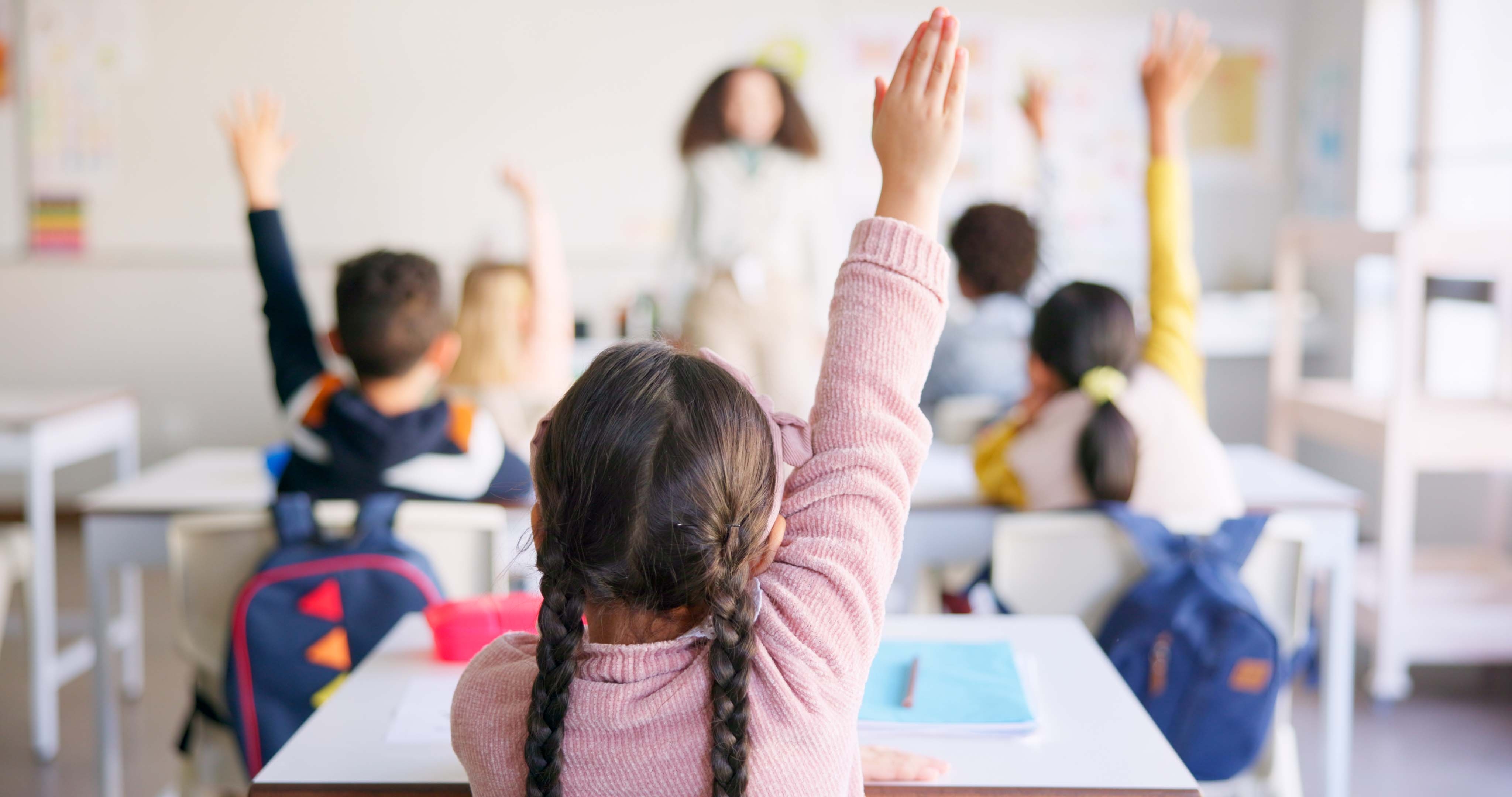Children in a classroom raising their hands to answer a question, with a teacher visible at the front of the class