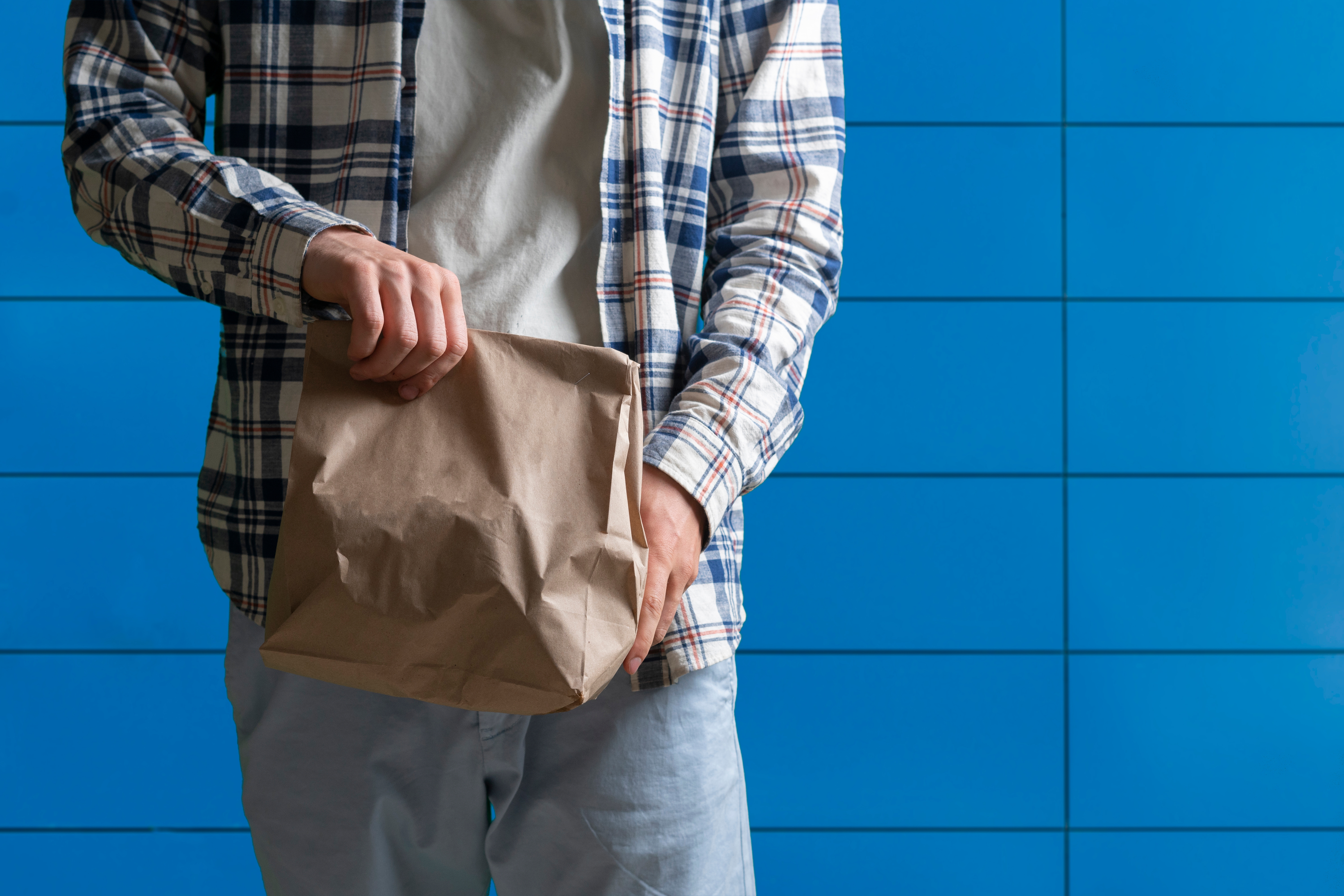 Person in casual clothes holding a crumpled paper bag, standing against a tiled background