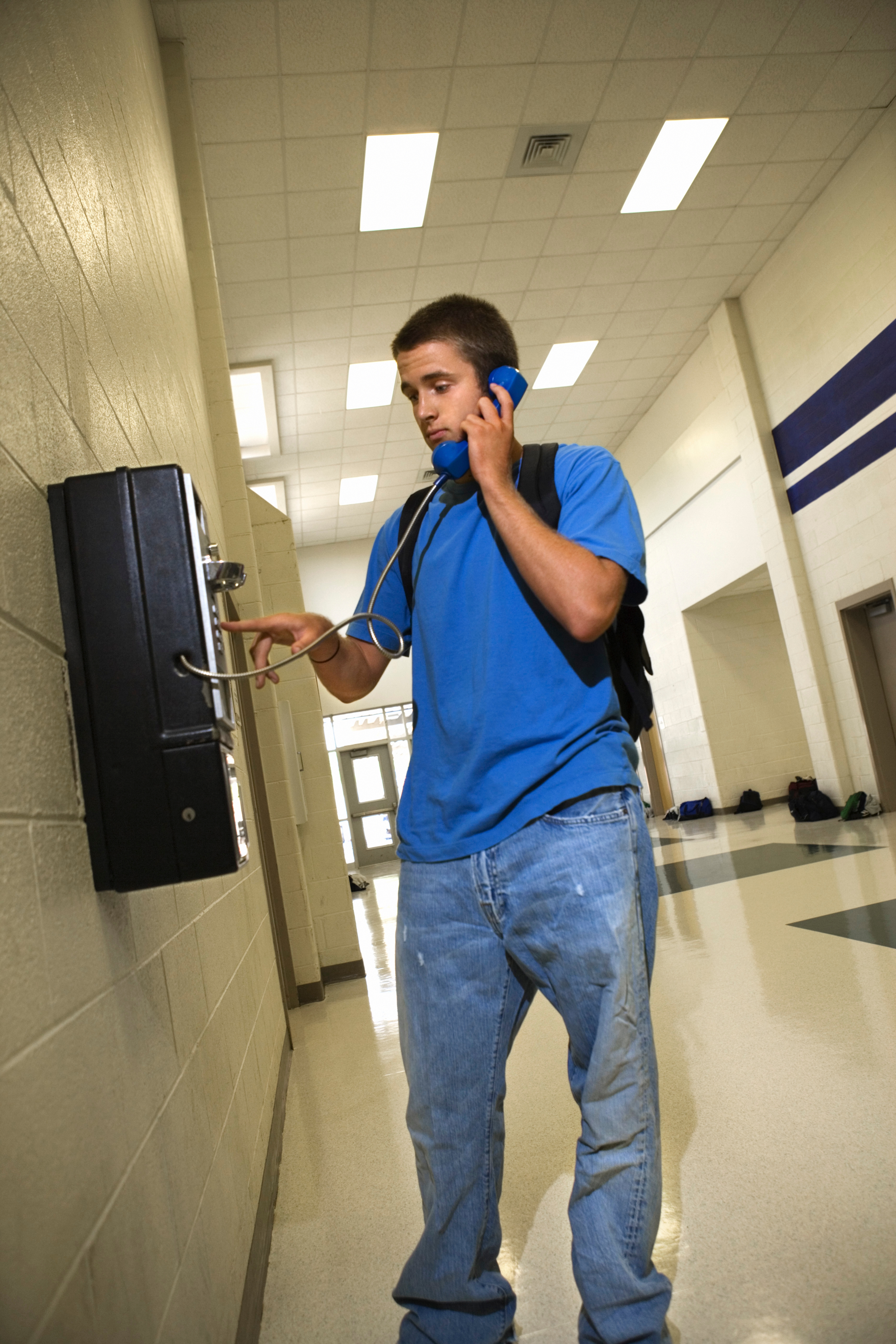Person in casual blue attire and jeans using a wall-mounted phone with a cord, in a hallway setting