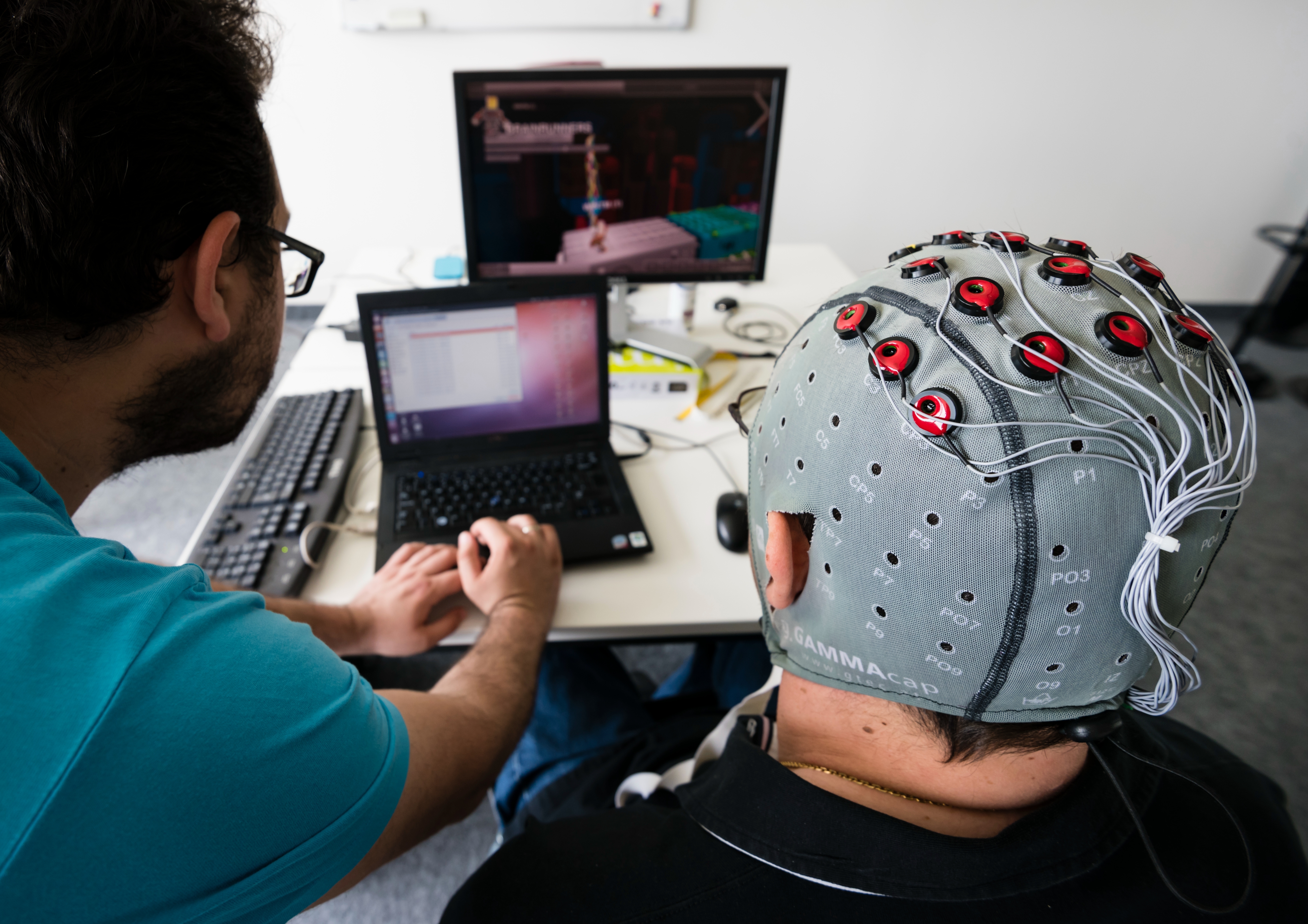 A person wearing an EEG cap sits at a desk viewing a laptop screen, collaborating with another individual observing the same screen