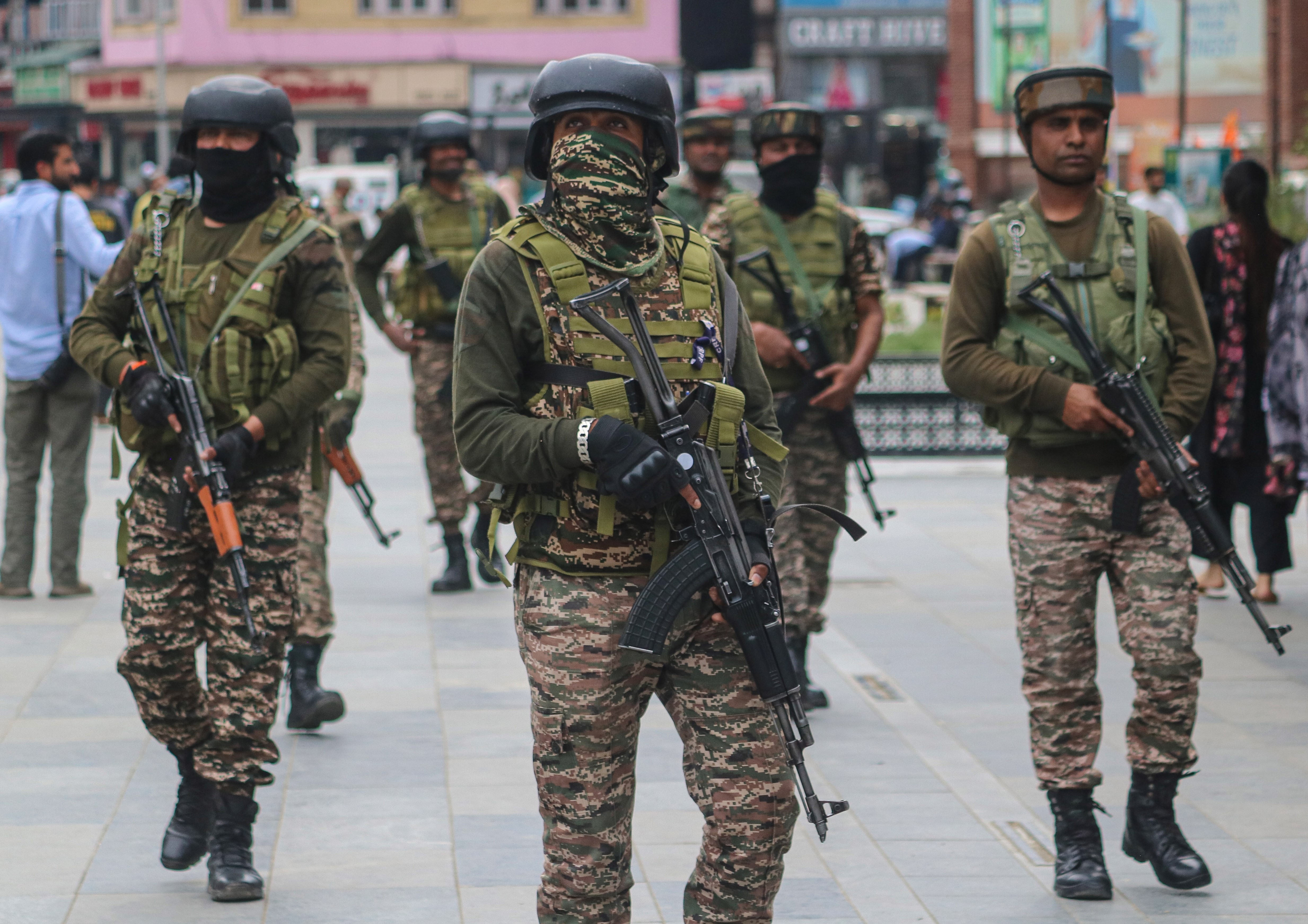 Soldiers in camouflage uniforms and helmets patrol a street, carrying rifles, with buildings visible in the background