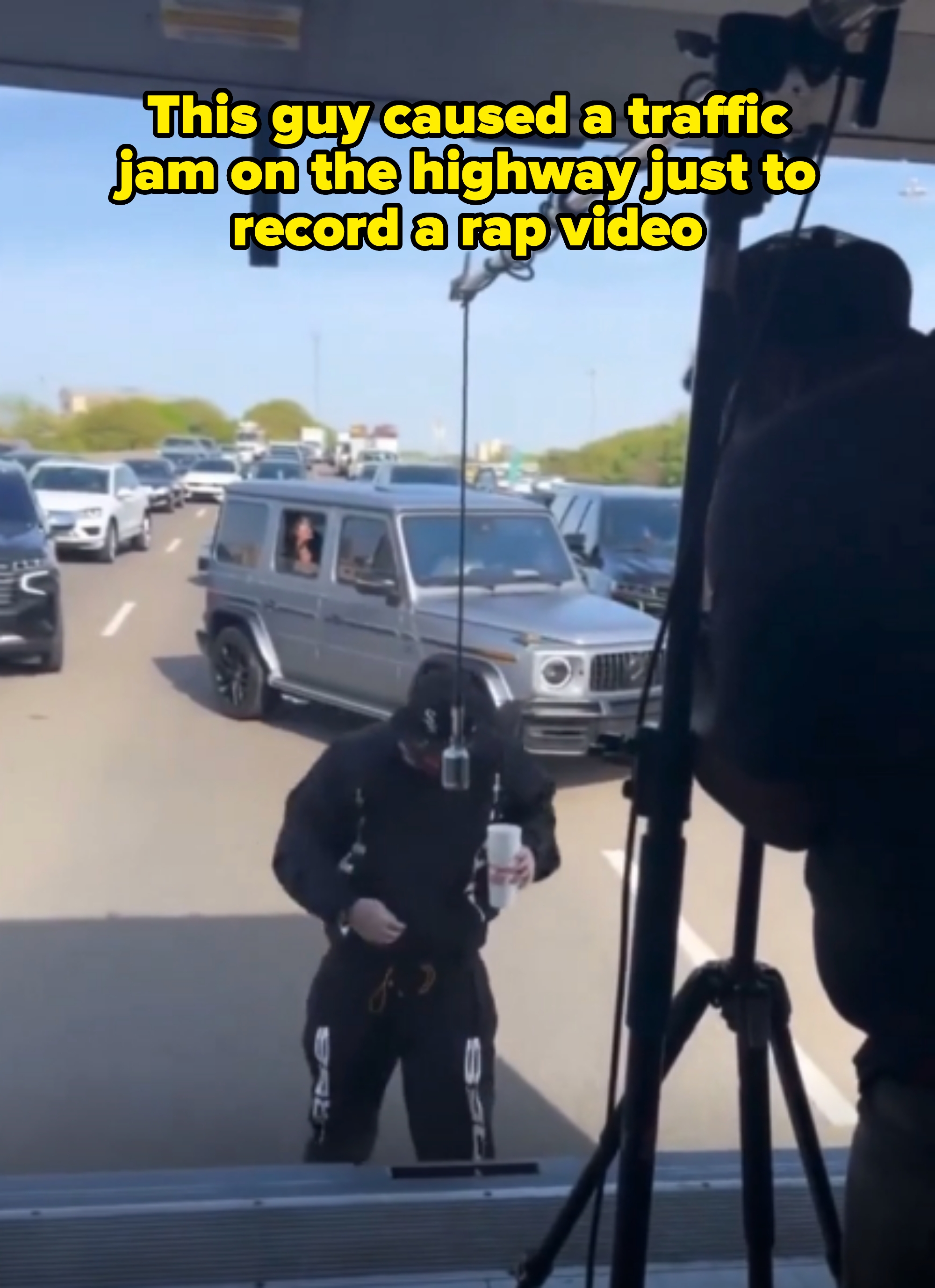 Person wearing streetwear, holding a drink, standing on a busy highway with crew and equipment visible. Caption: &quot;This guy caused a traffic jam on the highway just to record a rap video&quot;