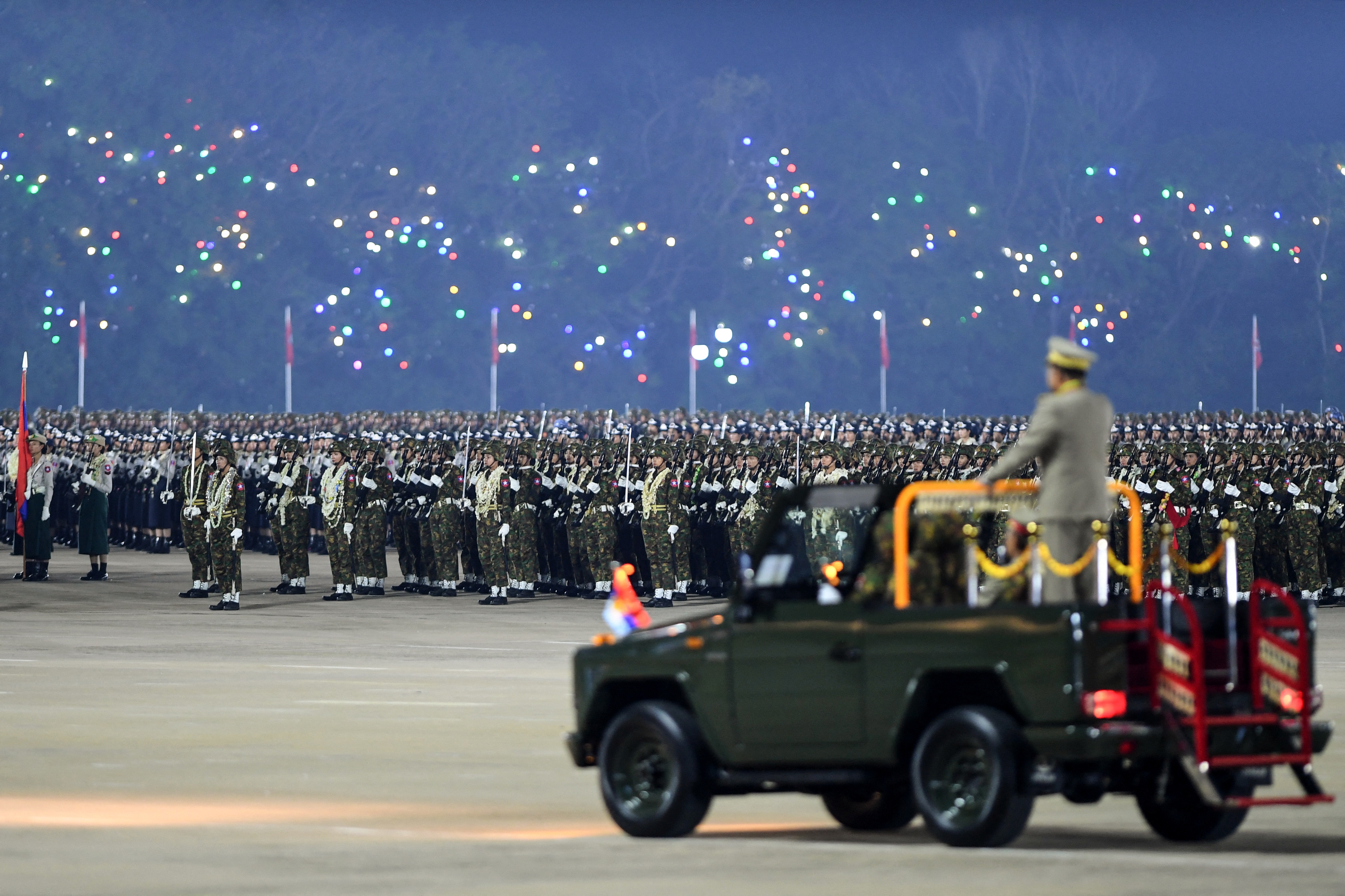 A large military parade is observed by an officer in a jeep, with hundreds of soldiers standing in formation and colorful lights in the background