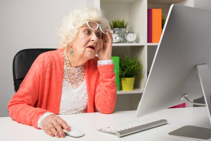 Elderly woman in white lace attire and coral cardigan, wearing glasses, looking intently at a computer screen in a modern home office
