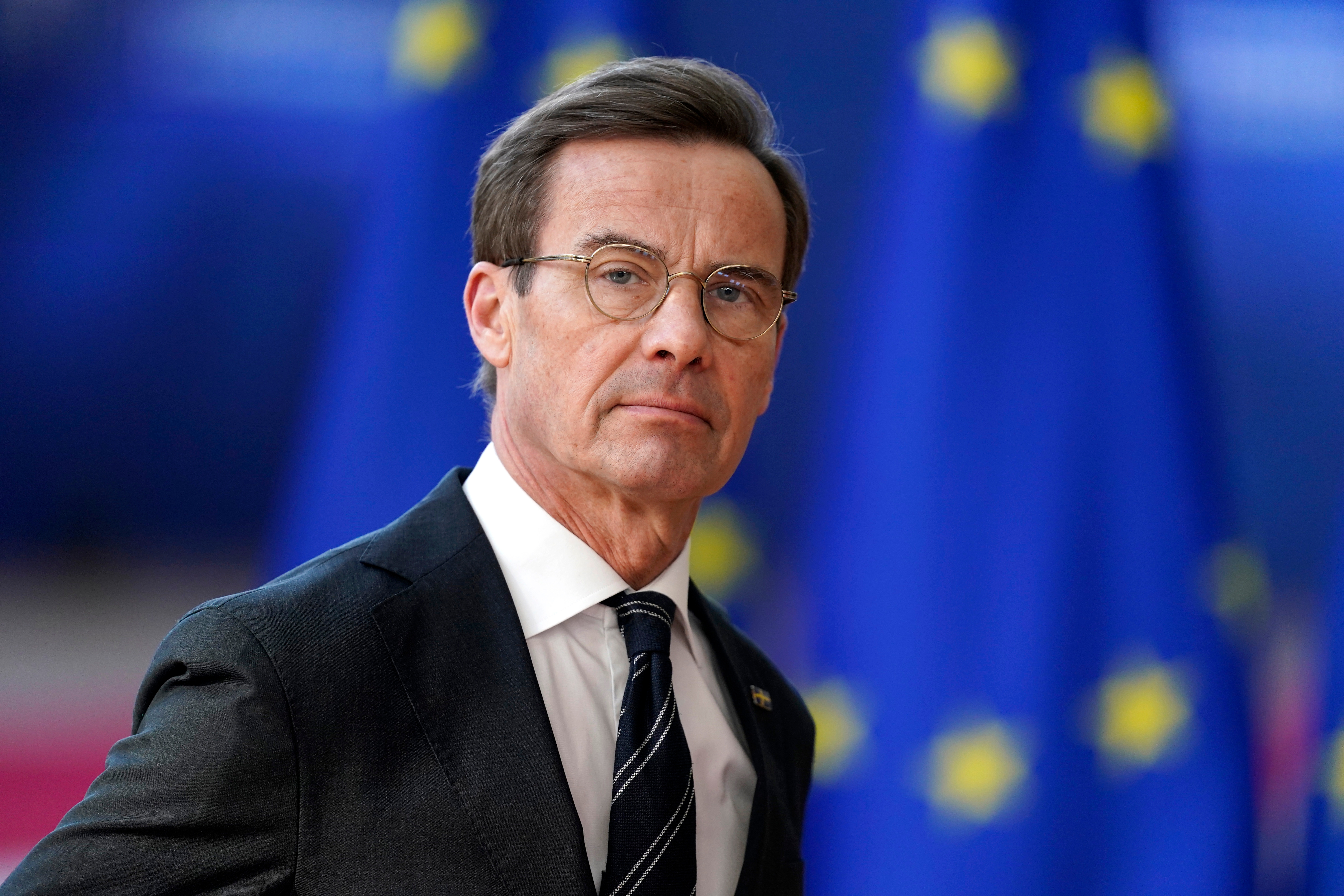 Man in suit and tie stands in front of EU flags, wearing glasses with a focused expression
