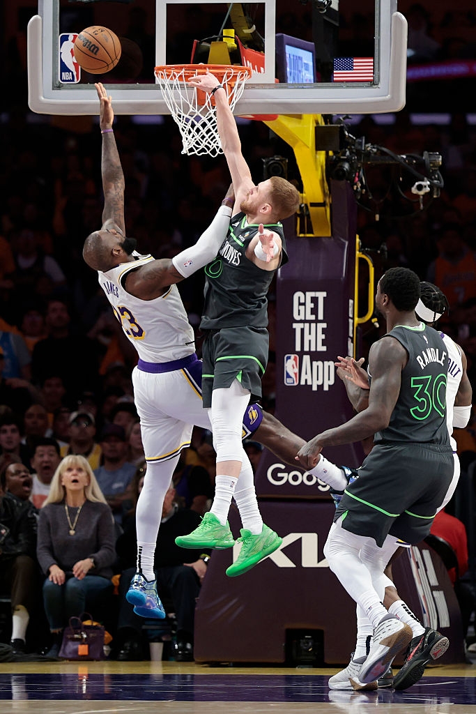 Basketball player attempts a layup while being blocked by a defender during an NBA game. Spectators and other players are visible in the background