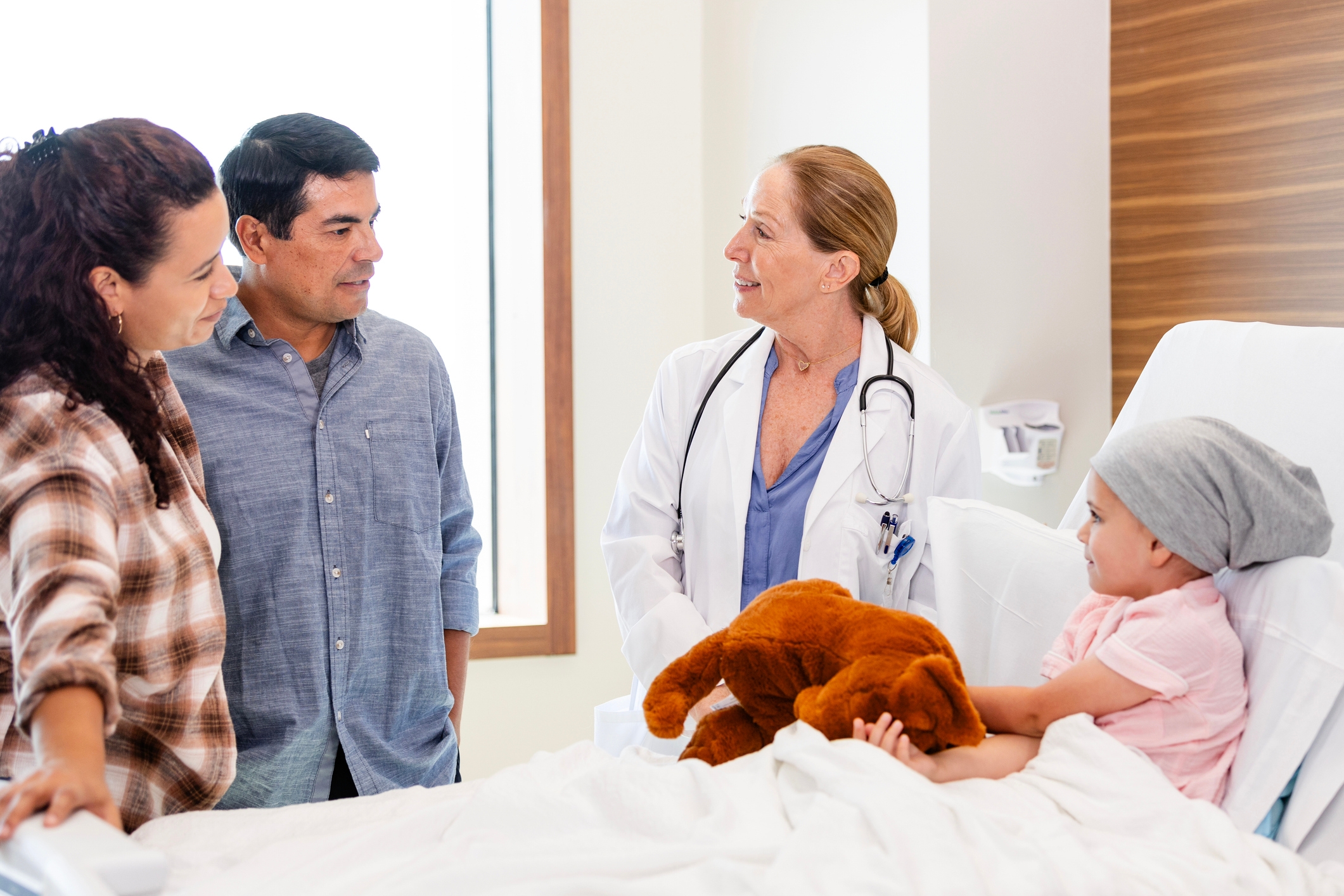 Doctor talks to parents while a child with a headscarf sits in a hospital bed holding a teddy bear, conveying a sense of care and support